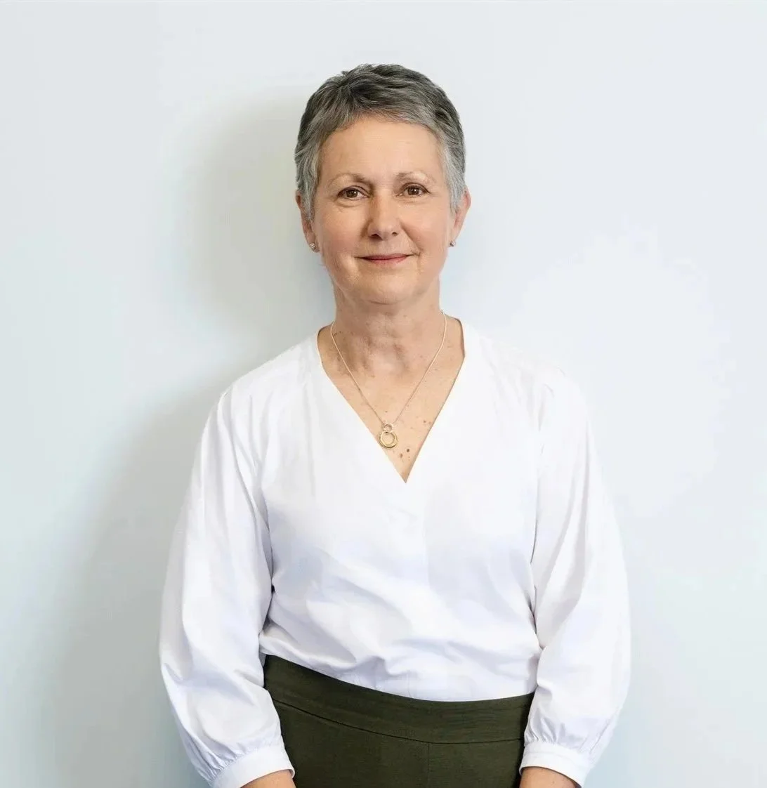 A middle-aged woman with short gray hair, wearing a white blouse and dark skirt, standing against a plain white background.
