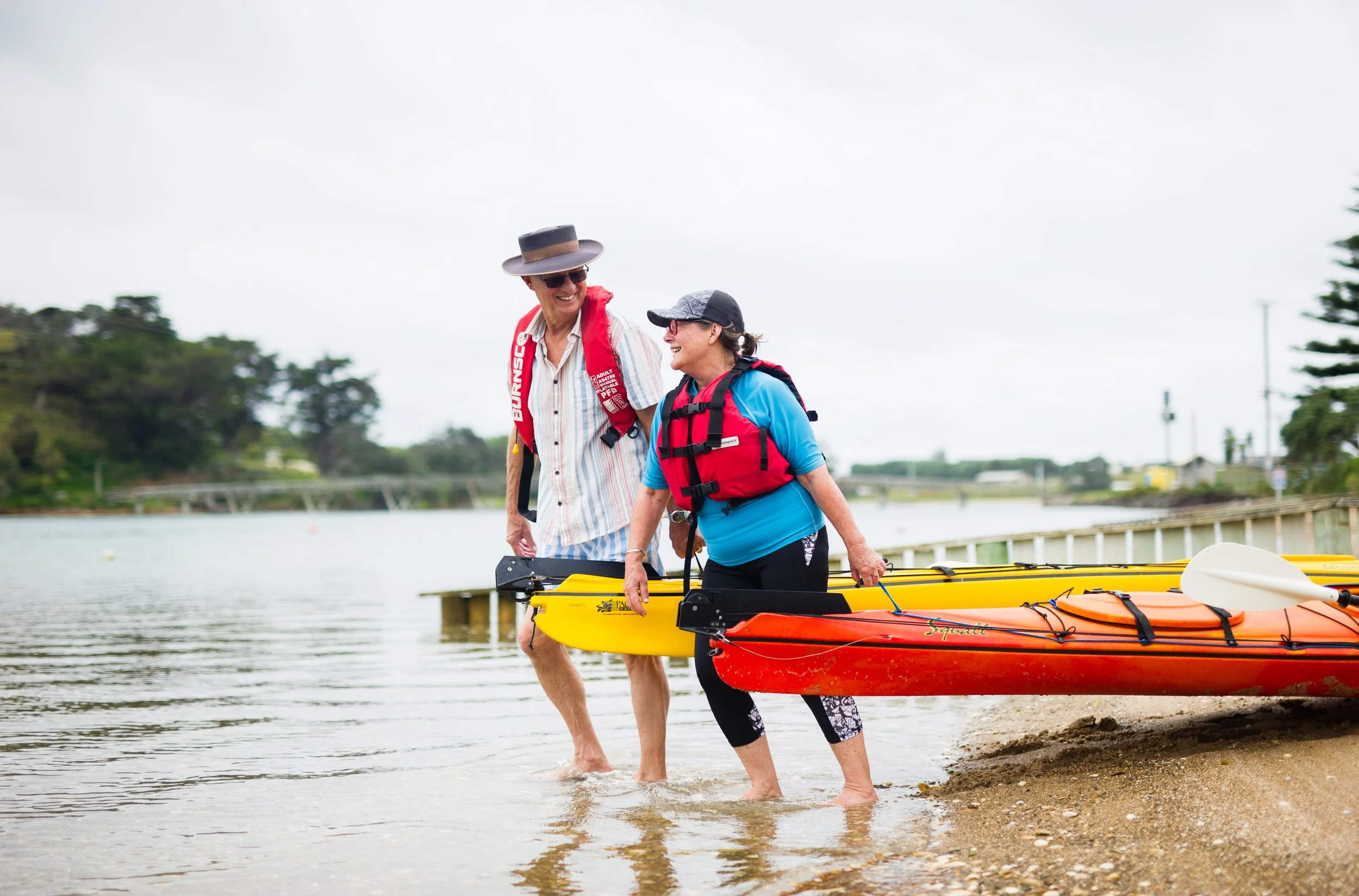 Two women in life jackets, one in a hat and the other wearing a cap, walking into the water with their kayaks on a sandy shore, smiling and talking.