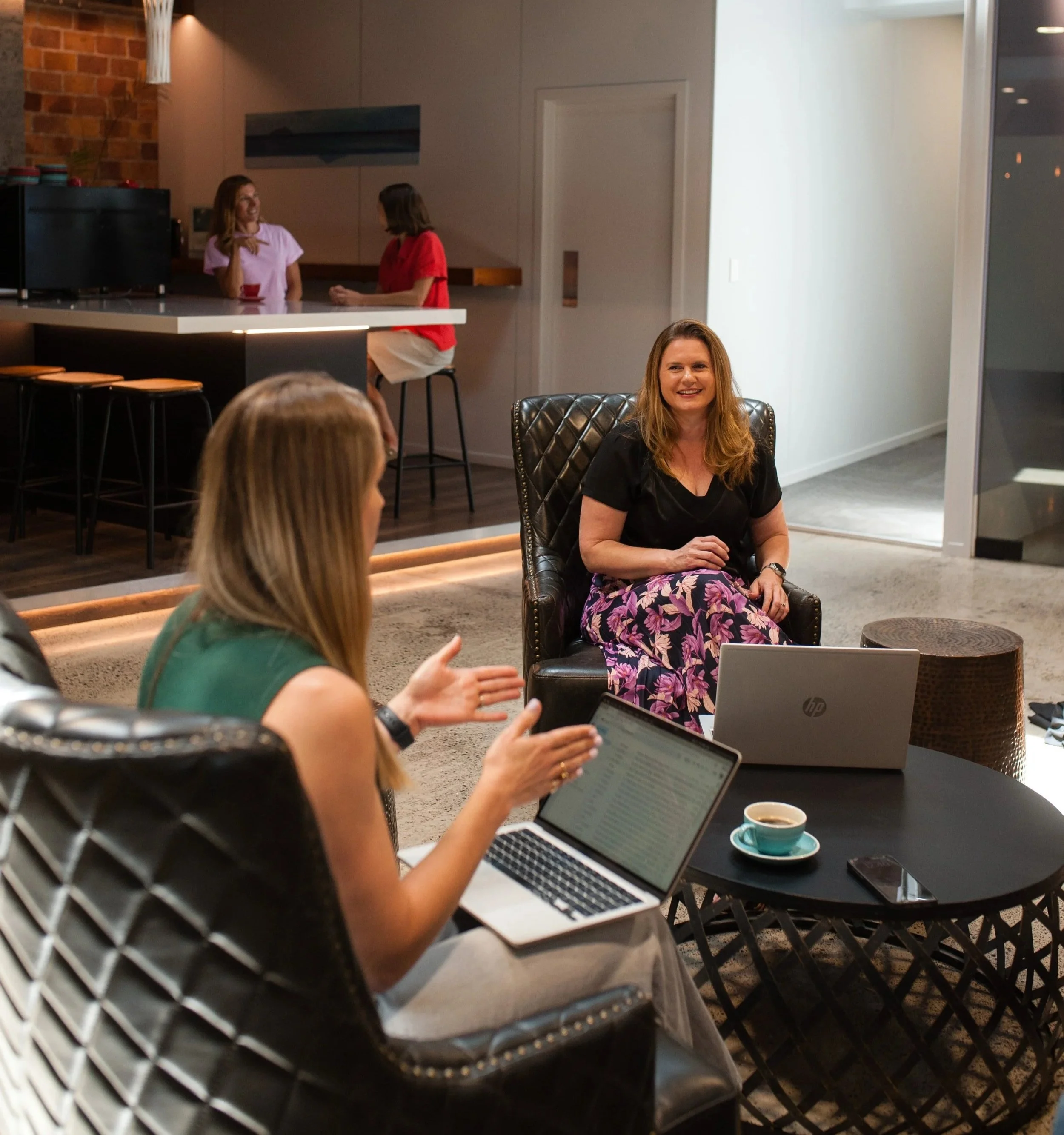 Group of women having a discussion in a modern living room with laptops and coffee, with two women talking at a kitchen counter in the background.