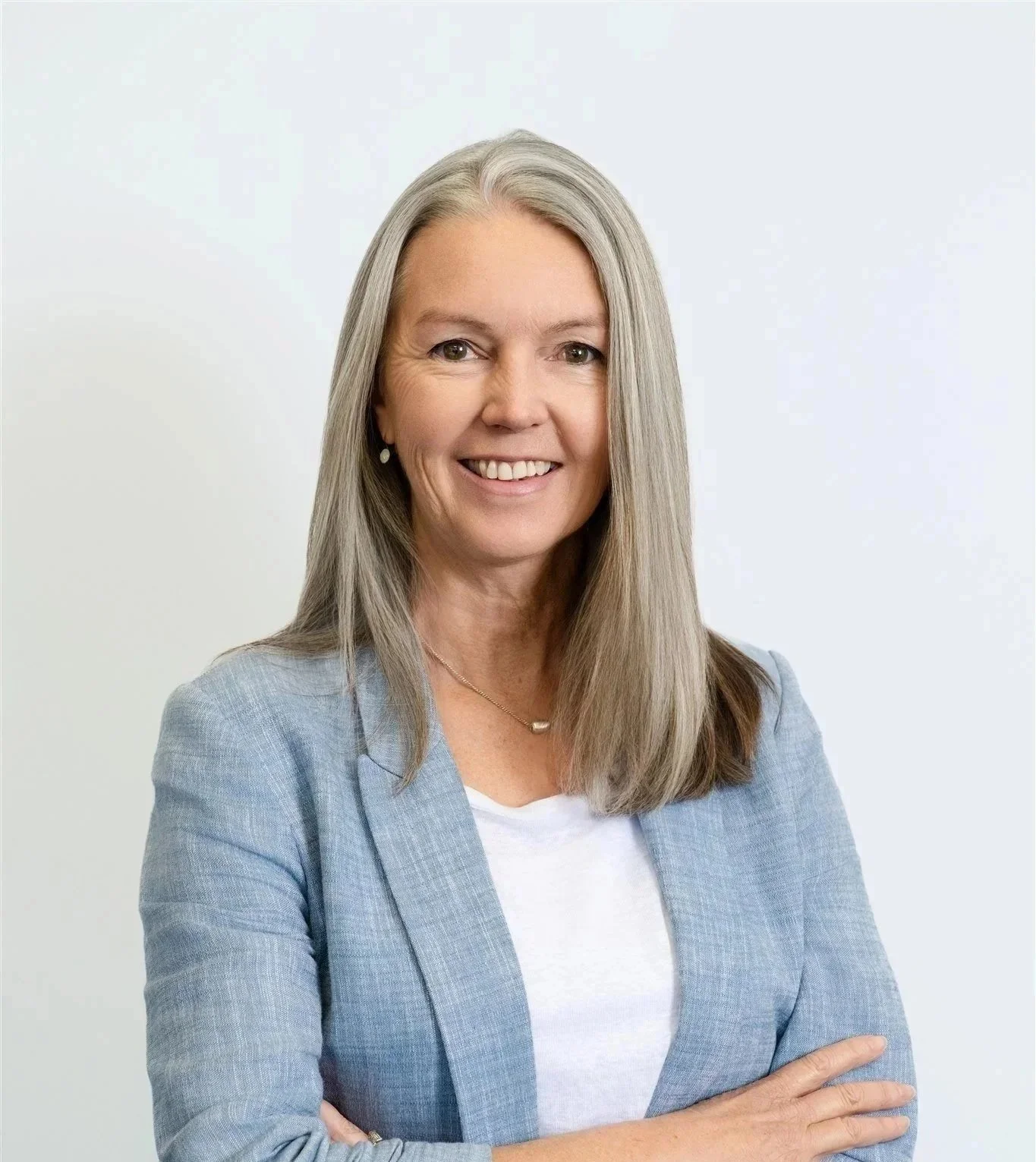 A middle-aged woman with long gray hair, smiling, wearing a light blue blazer and a white top, standing against a plain white background.