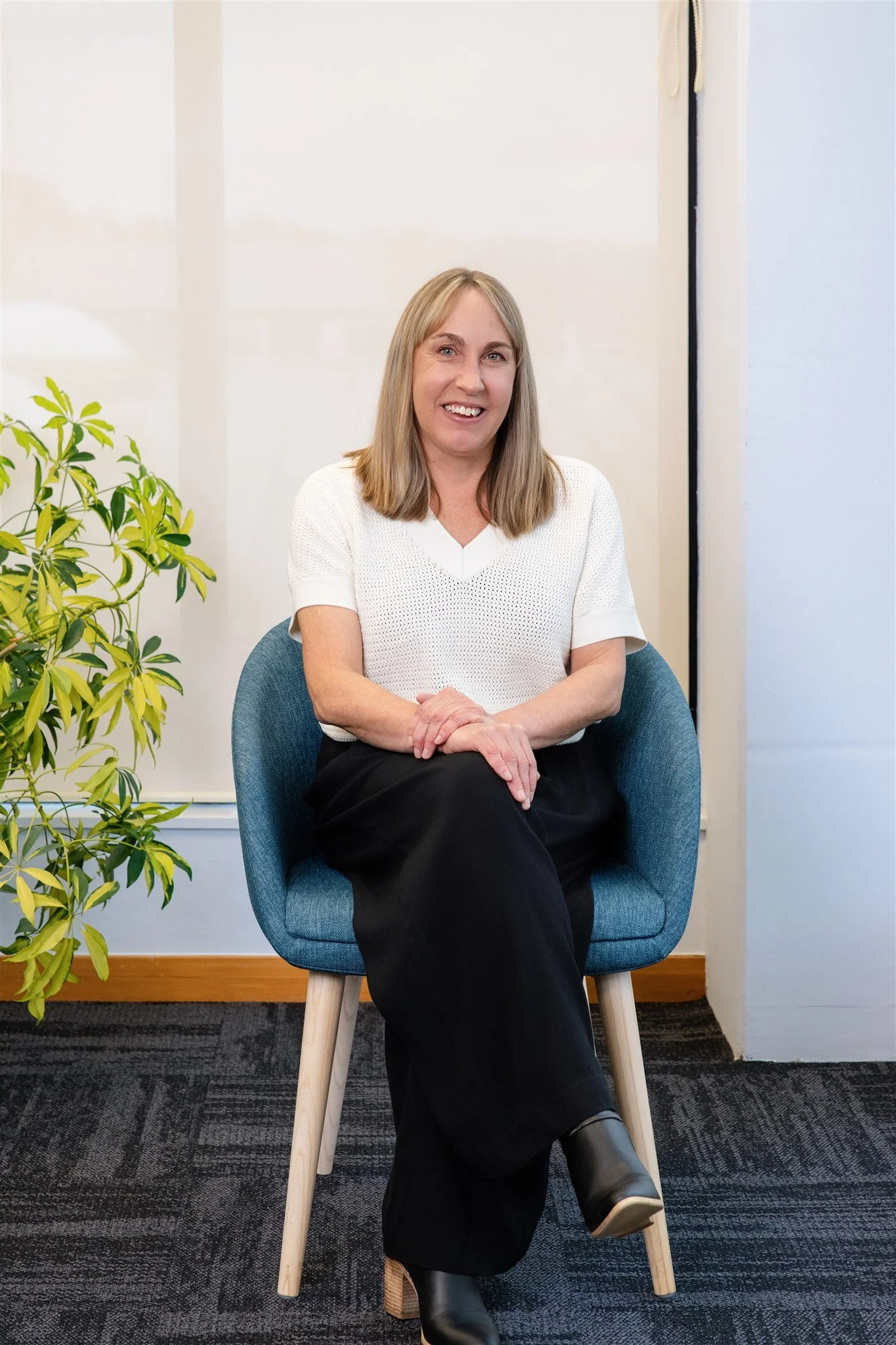 A woman with shoulder-length blonde hair smiling, sitting on a blue chair with her hands folded in her lap. She is wearing a white short-sleeve top and black pants, in an indoor setting with a potted plant nearby.