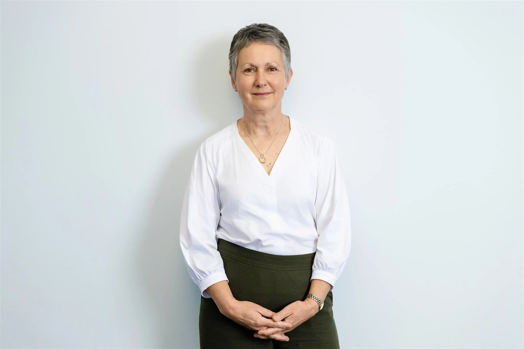 A professional woman with short gray hair, wearing a white blouse and dark pants, standing against a plain white wall.