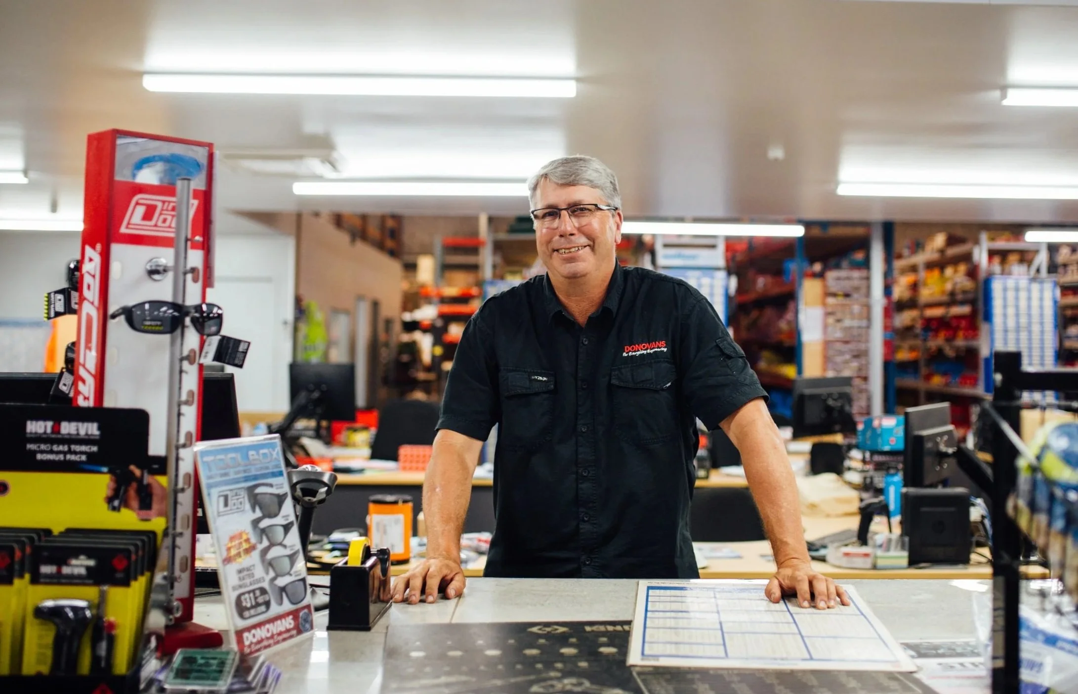A man with gray hair and glasses, wearing a black Donovans shirt, stands behind a counter in a hardware or retail store, smiling at the camera.