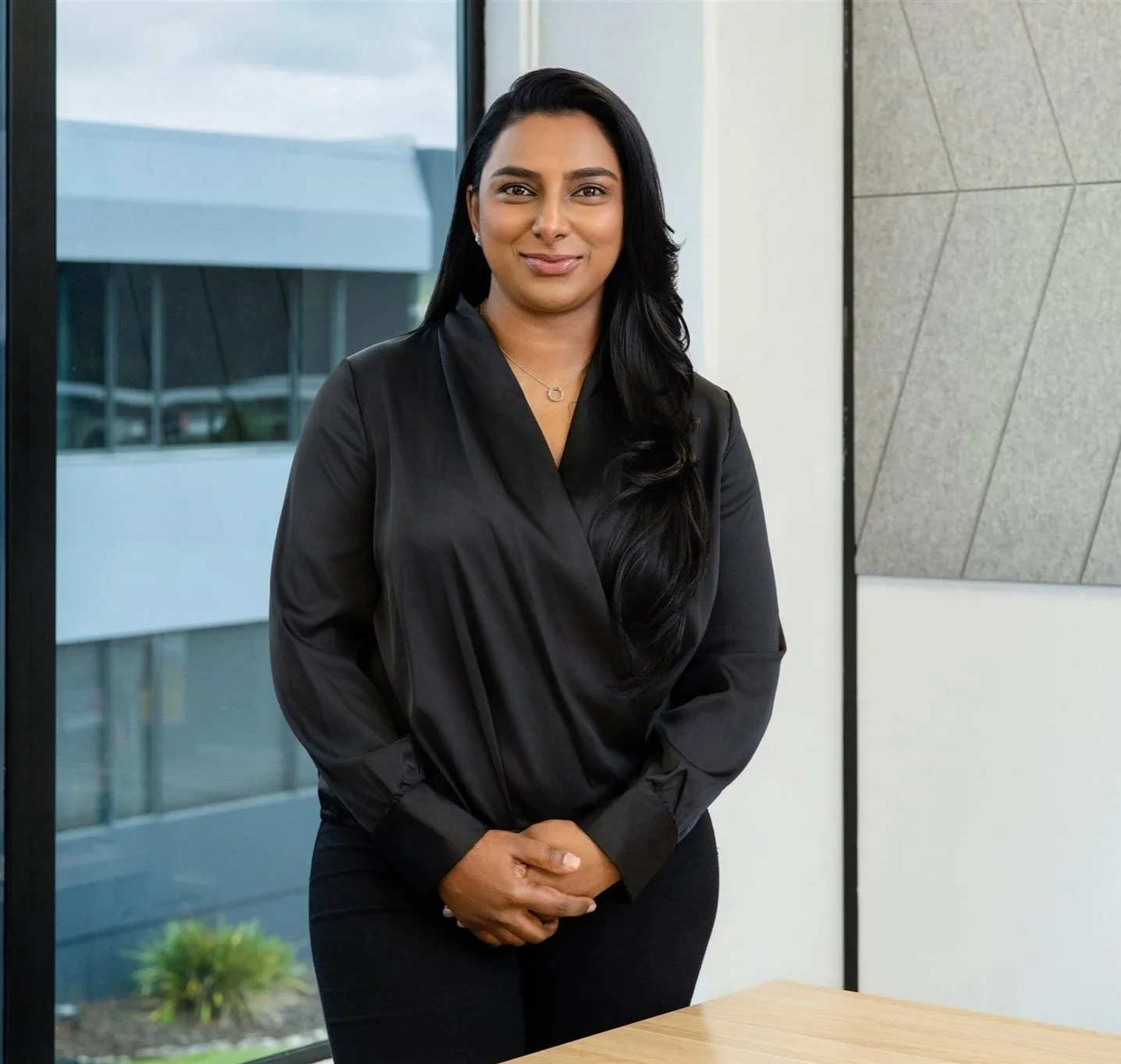 A woman with long black hair wearing a black blouse and black pants, standing indoors near a window and wall, smiling at the camera.