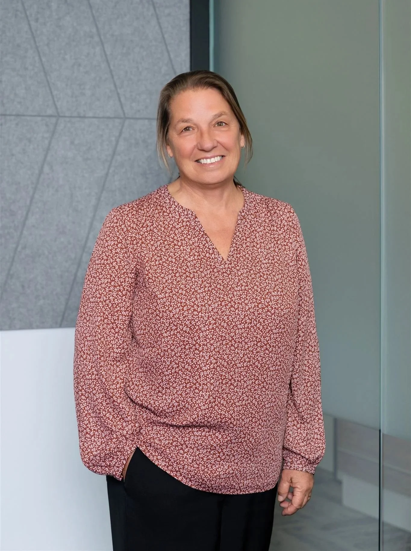 A smiling woman with shoulder-length hair wearing a red patterned blouse and black pants, standing indoors next to a textured gray wall and a glass partition.