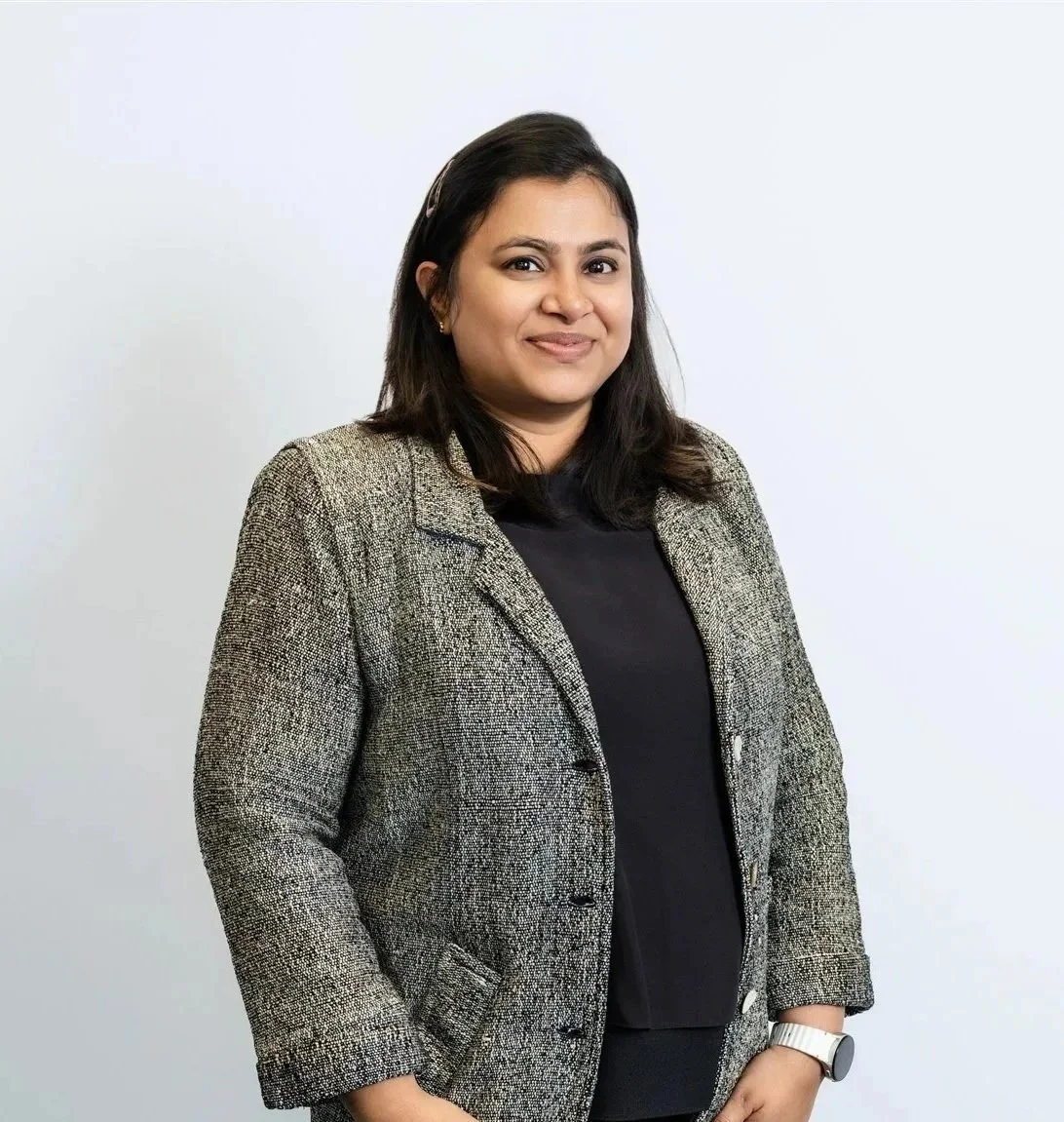 A woman with black hair, wearing a black shirt and a gray blazer, standing against a plain white background.