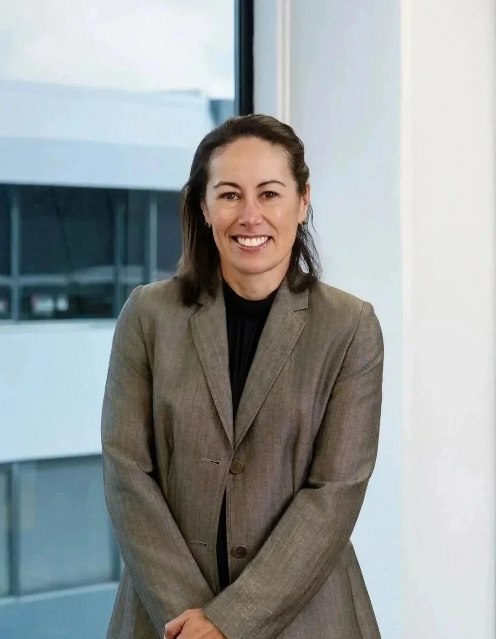 A woman in a business suit smiling, standing indoors near a window with a city building in the background.