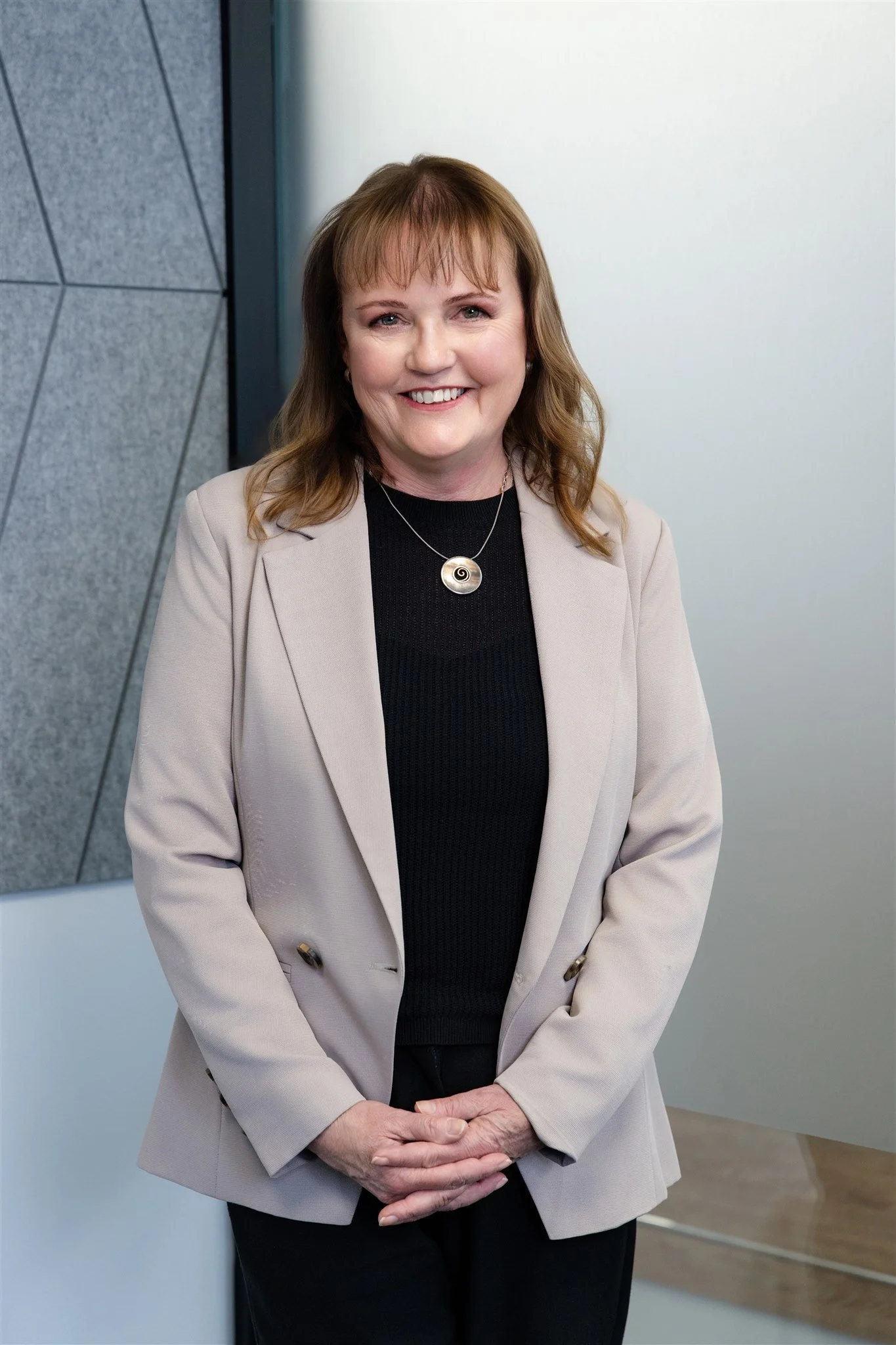 A smiling woman with shoulder-length brown hair, wearing a beige blazer over a black top, standing indoors against a modern wall background.