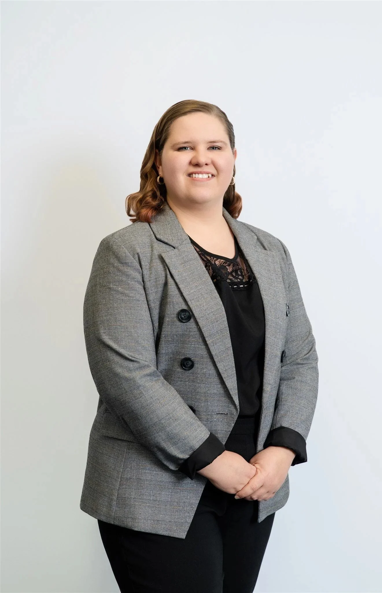 A woman with shoulder-length brown hair, wearing a gray blazer over a black lace blouse, smiling against a plain white background.