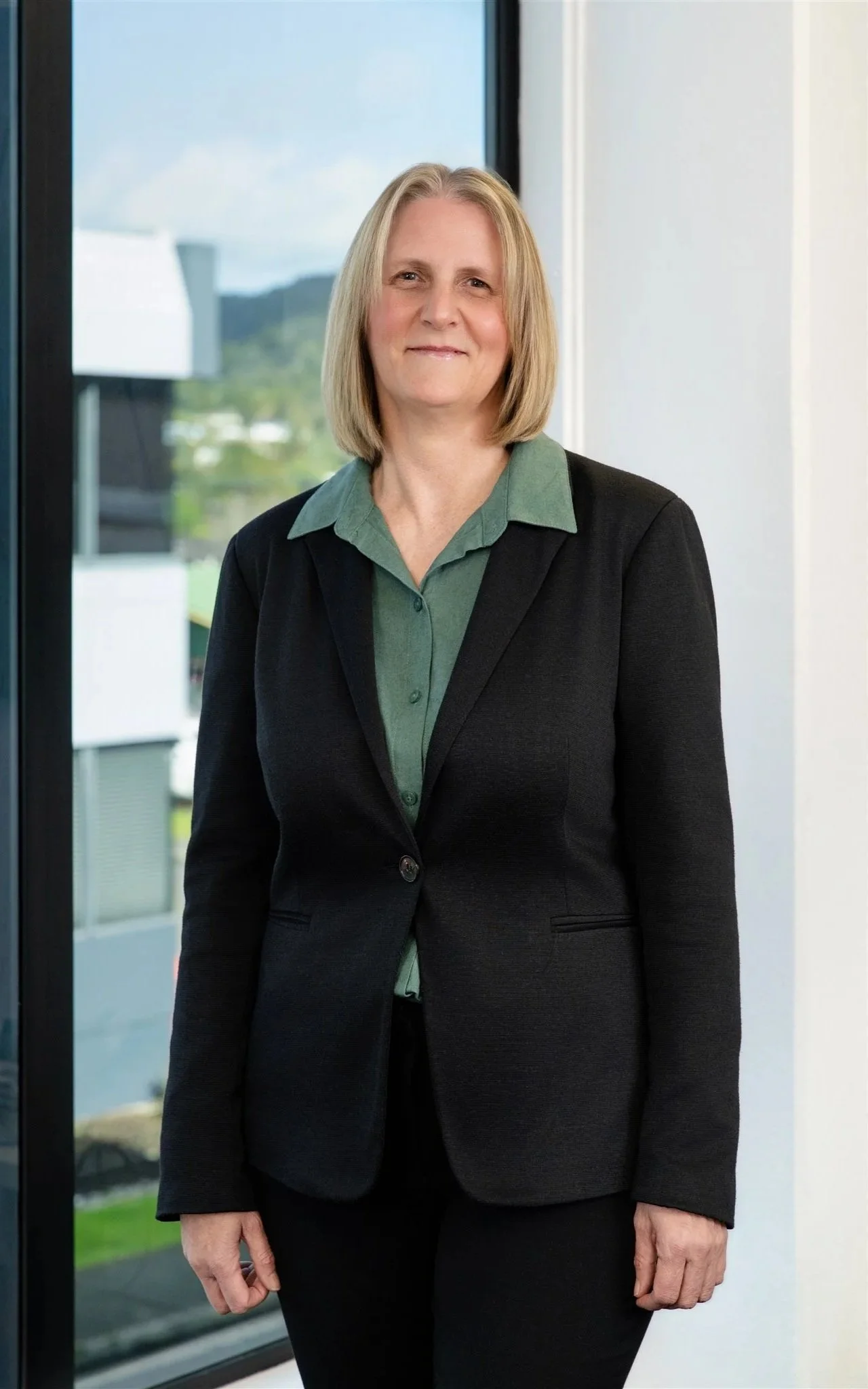 A professional woman standing indoors in front of a window with a cityscape view, wearing a black blazer over a green collared shirt.