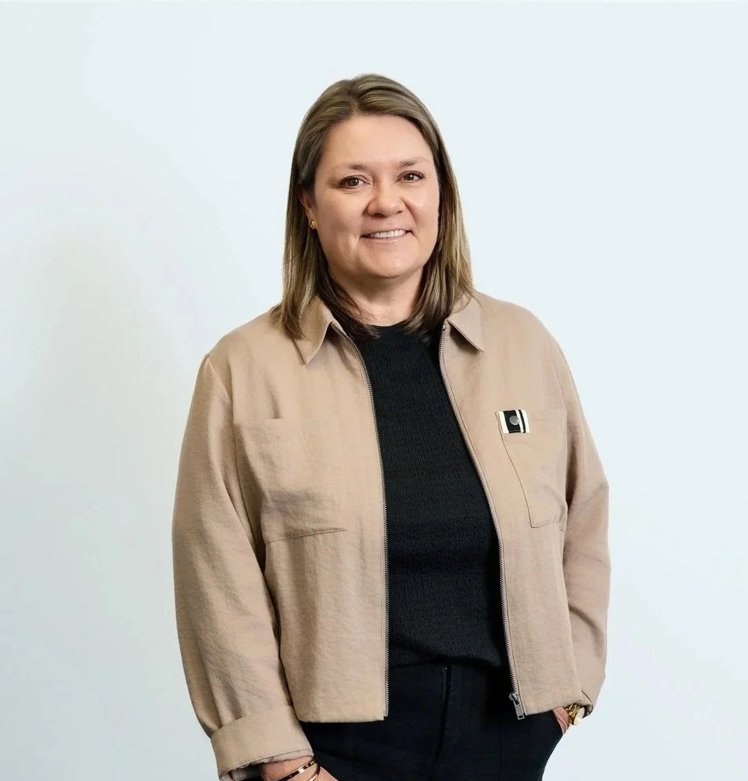A woman with shoulder-length brown hair smiling, wearing a beige jacket over a black top, standing against a plain light blue background.