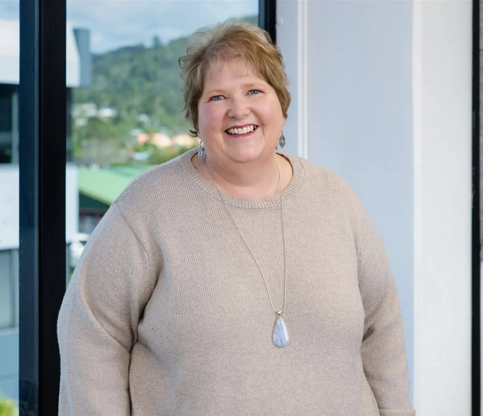 Smiling woman with short, light brown hair standing indoors near a large window, wearing a beige sweater and a long necklace with a large pendant.