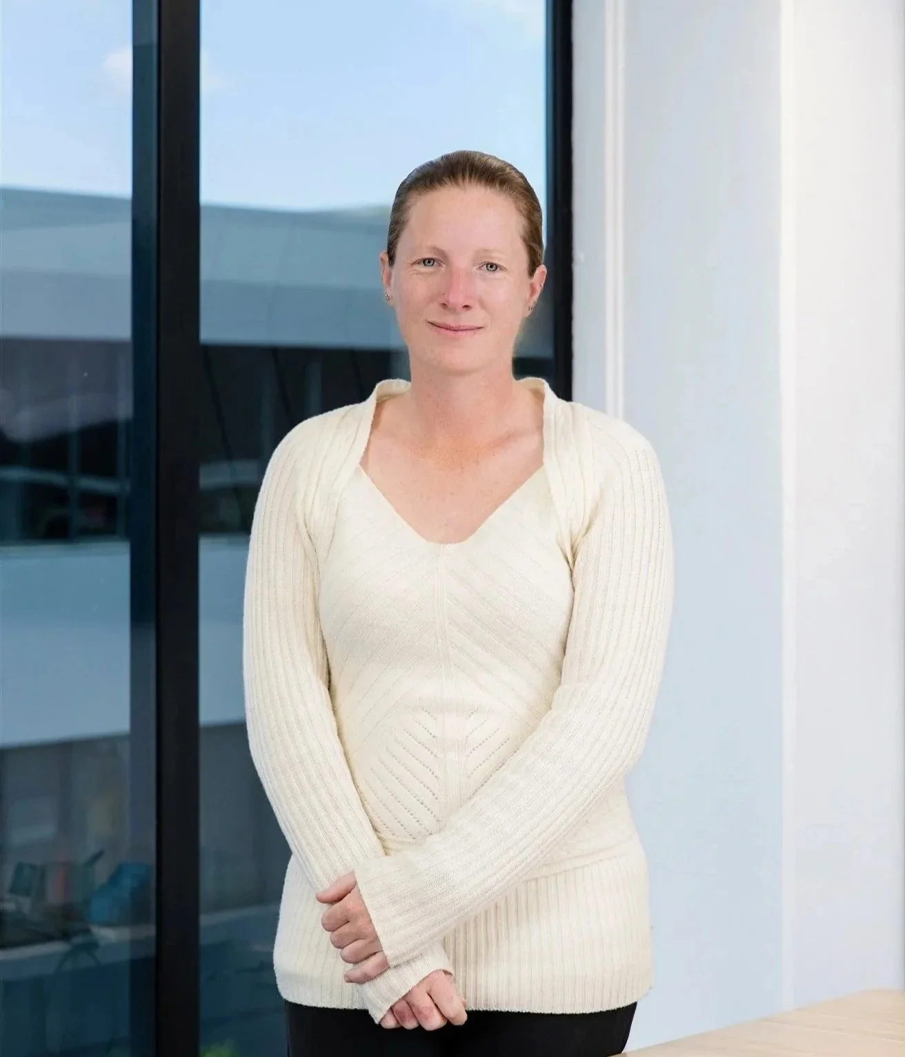 A woman with short brown hair standing near a window, wearing a cream-colored sweater, smiling at the camera.