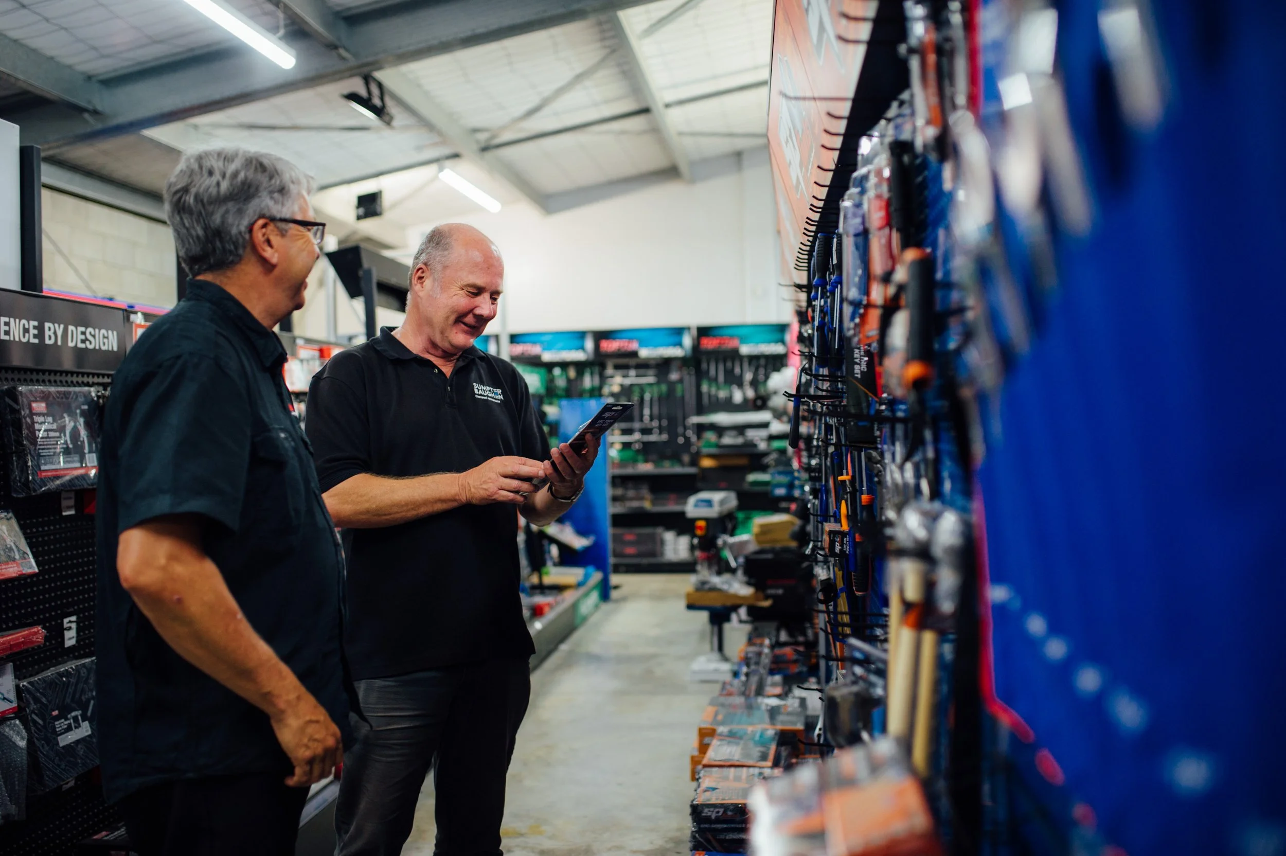 Two men shopping in a hardware store, looking at a display of tools on the wall.
