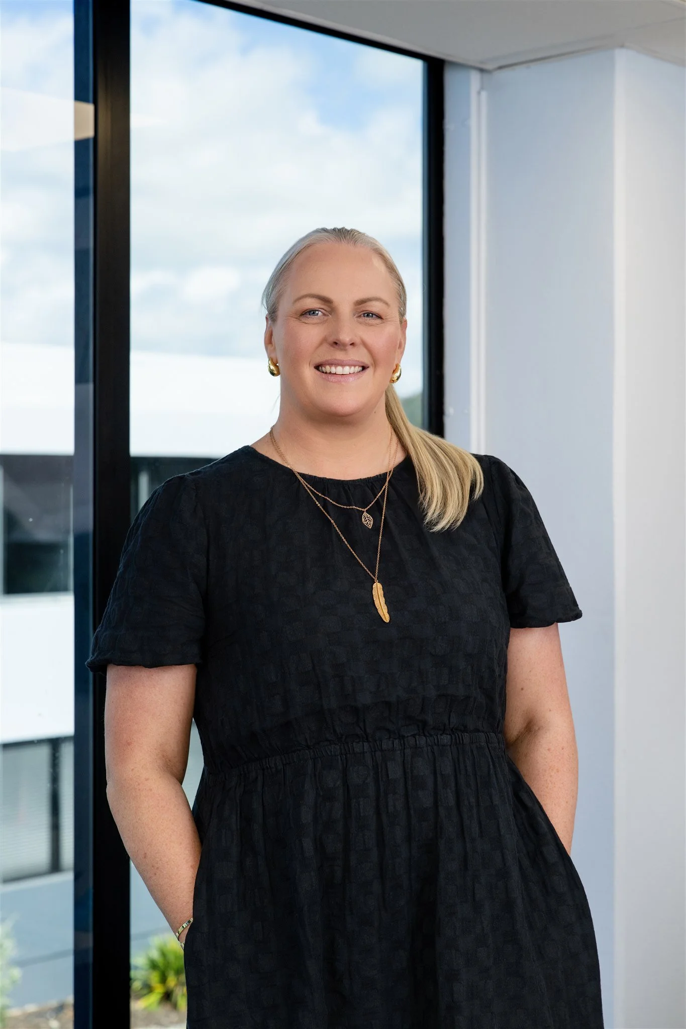 A woman standing indoors near a large window, smiling at the camera, with a view of blue skies and clouds outside.