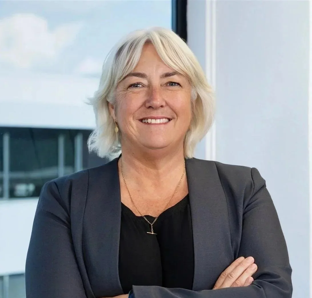 A professional woman with short gray hair, wearing a dark blazer and a black top, standing with her arms crossed in an office setting, smiling at the camera.