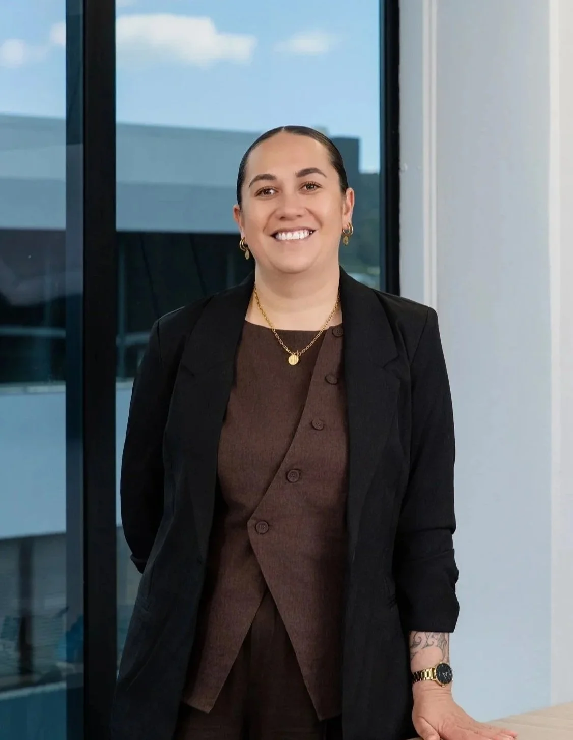 A woman standing indoors next to a glass wall, smiling at the camera, wearing a black blazer over a brown shirt, gold jewelry, and a gold watch.