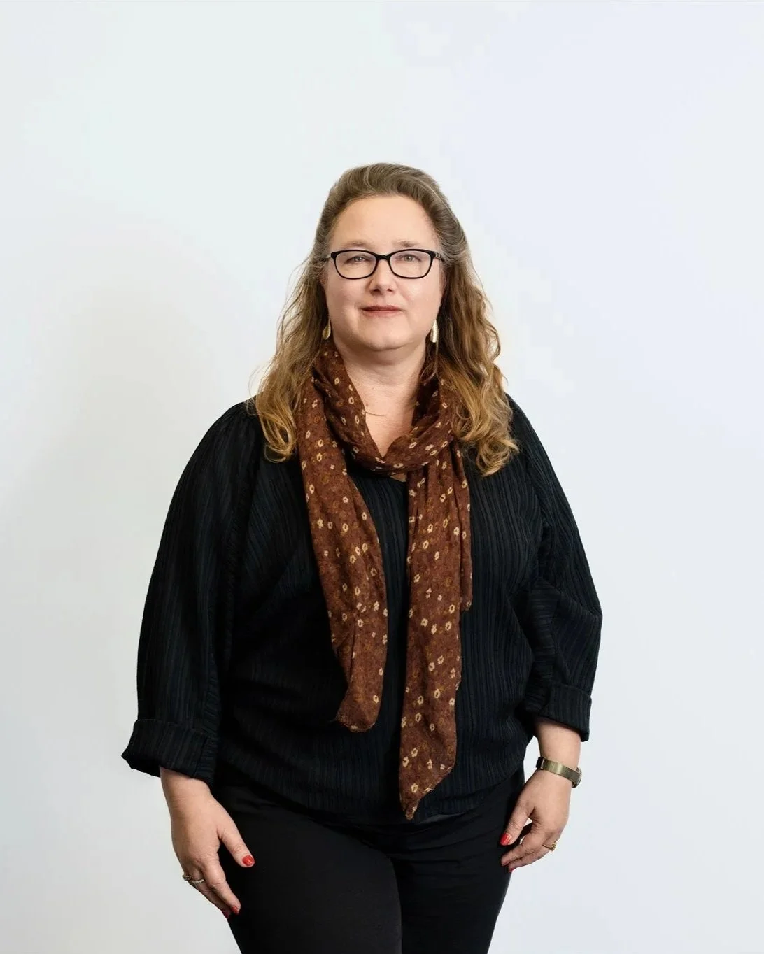 Portrait of a woman with glasses and wavy hair, wearing a black top, a brown scarf, and dark pants, standing against a plain white background.