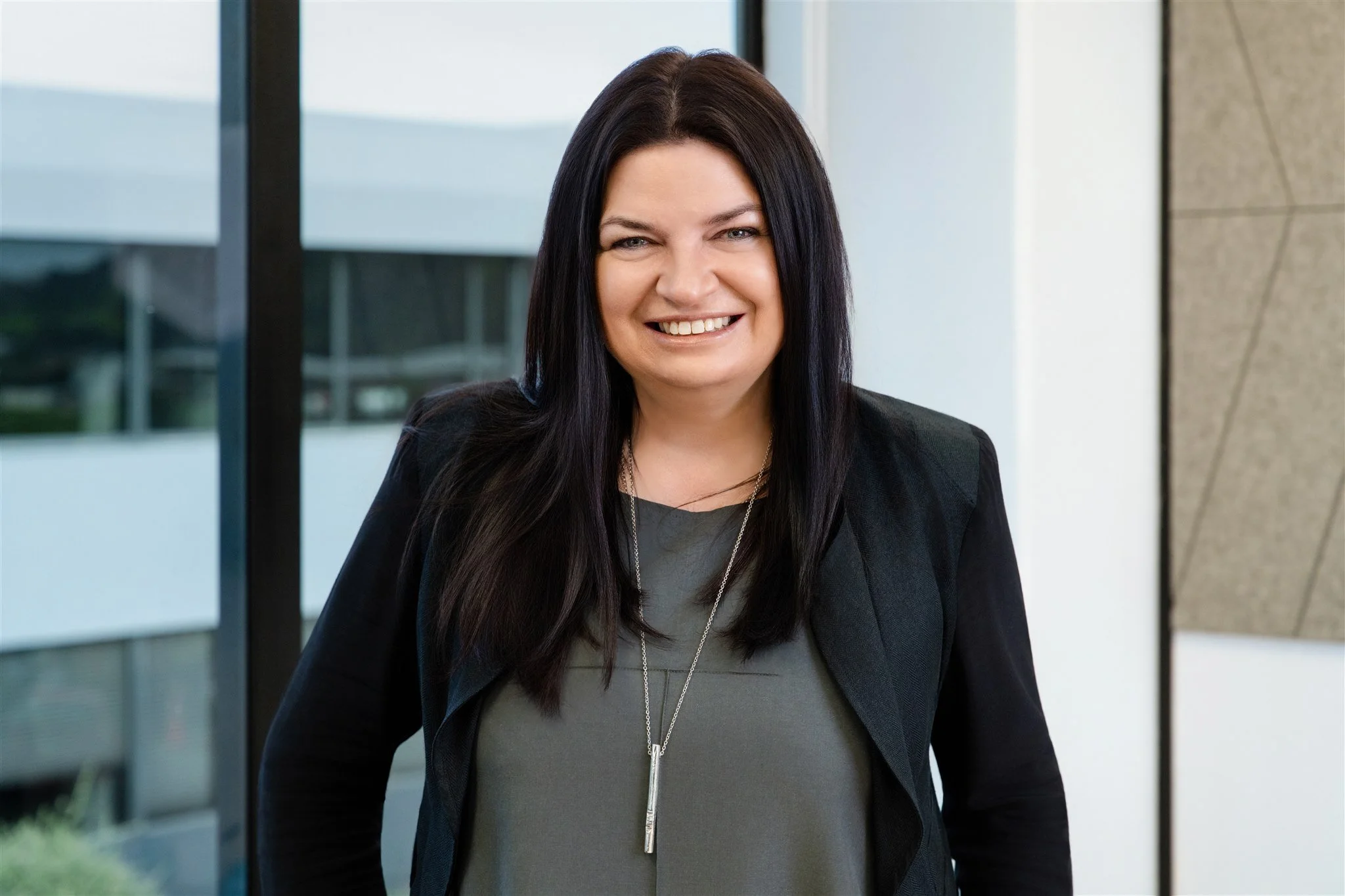 A woman with long dark hair and a gray top, wearing a black blazer and a silver necklace, smiling in an office setting with large windows.