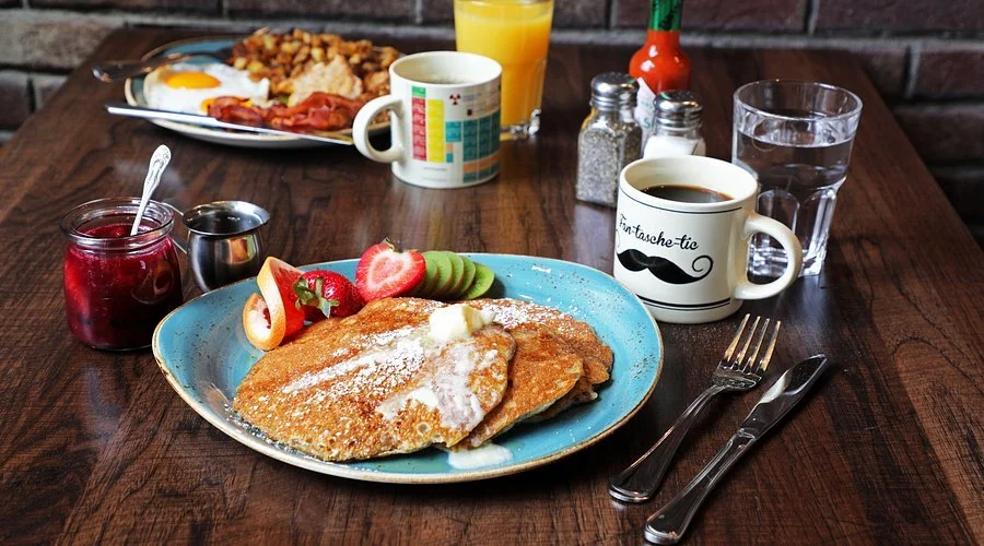 Breakfast with pancakes topped with powdered sugar and fruit, a plate of breakfast with eggs and potatoes, and various drinks including orange juice, coffee, water, and jam on a wooden table.