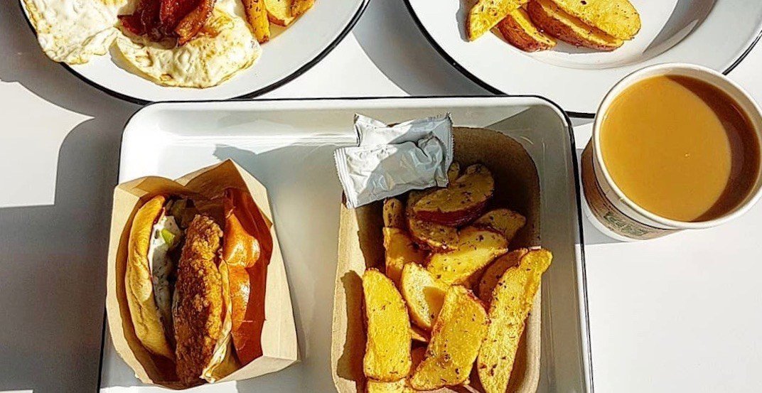 Fast food meal on white tray including a fried chicken sandwich, seasoned potato wedges, and a cup of orange juice. Additional plates with potato wedges and fried eggs with bacon are visible in the background.