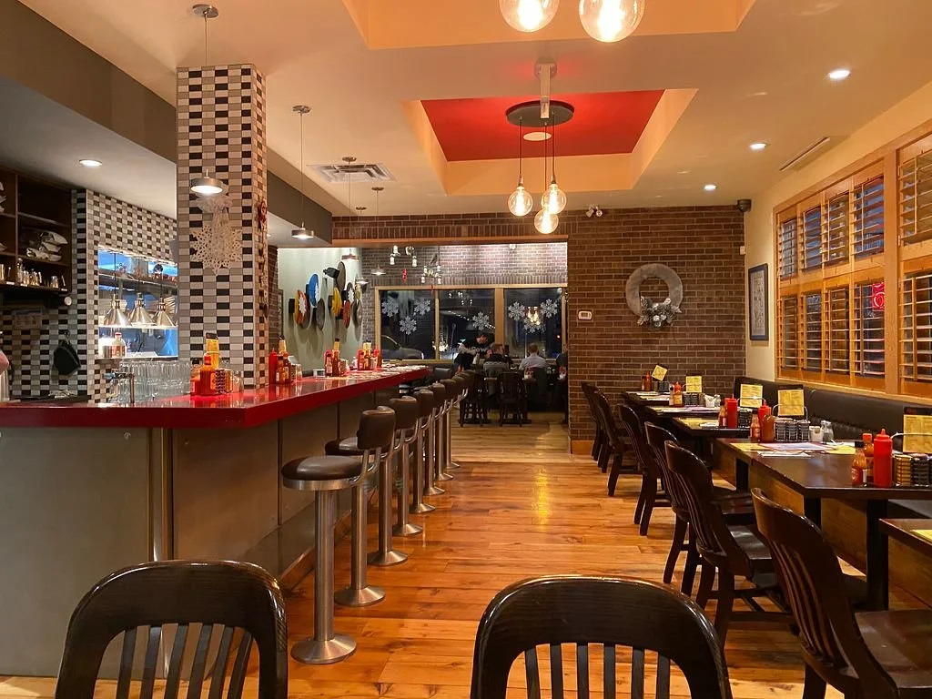 Interior of a cozy restaurant with a bar counter on the left, wooden flooring, tables with food menus and condiments, brick walls, and warm lighting.