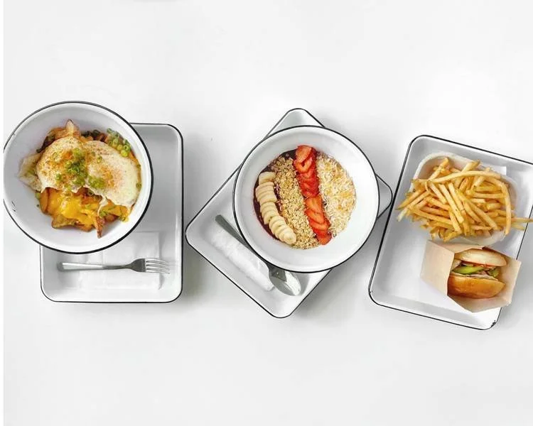 Overhead view of three plates with different meals: a bowl of shepherd's pie, a bowl with sliced bananas, strawberries, and granola, and a plate with French fries and a small burger in a paper tray on a white surface.