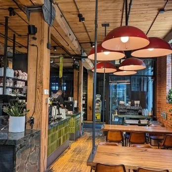 Interior of a cozy cafe with wooden beams, decorative red pendant lights, a counter area with coffee equipment, and several wooden tables with chairs.