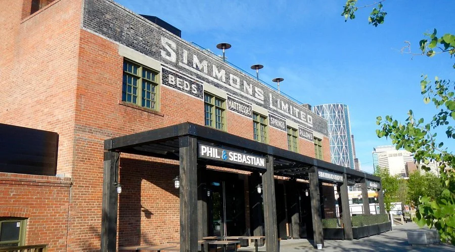 A brick building with a sign for Simmons Limited, displaying services like beds, matteresses, springs, pillows, and Villecoans. Below is a black awning with a sign reading 'Phil & Sebastian'.