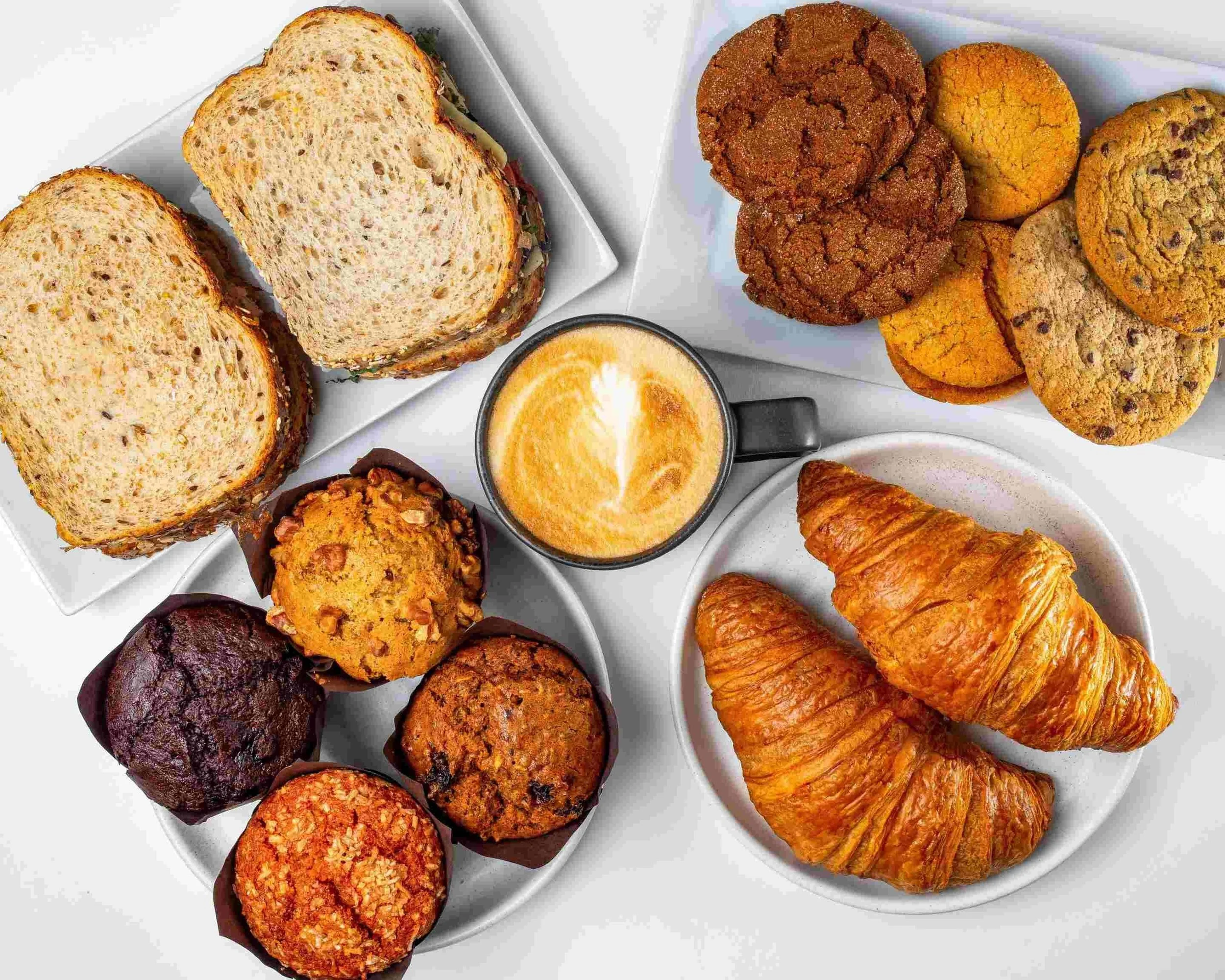 A breakfast spread with sliced bread, assorted cookies, muffins, croissants, and a cup of coffee.