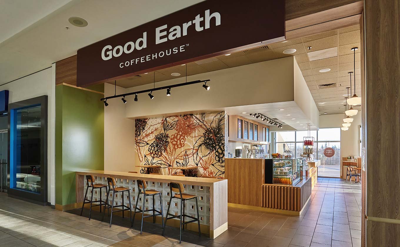 Interior of a coffee shop named Good Earth Coffeehouse, featuring a counter with barstools, a decorative wall, and seating area with large windows.