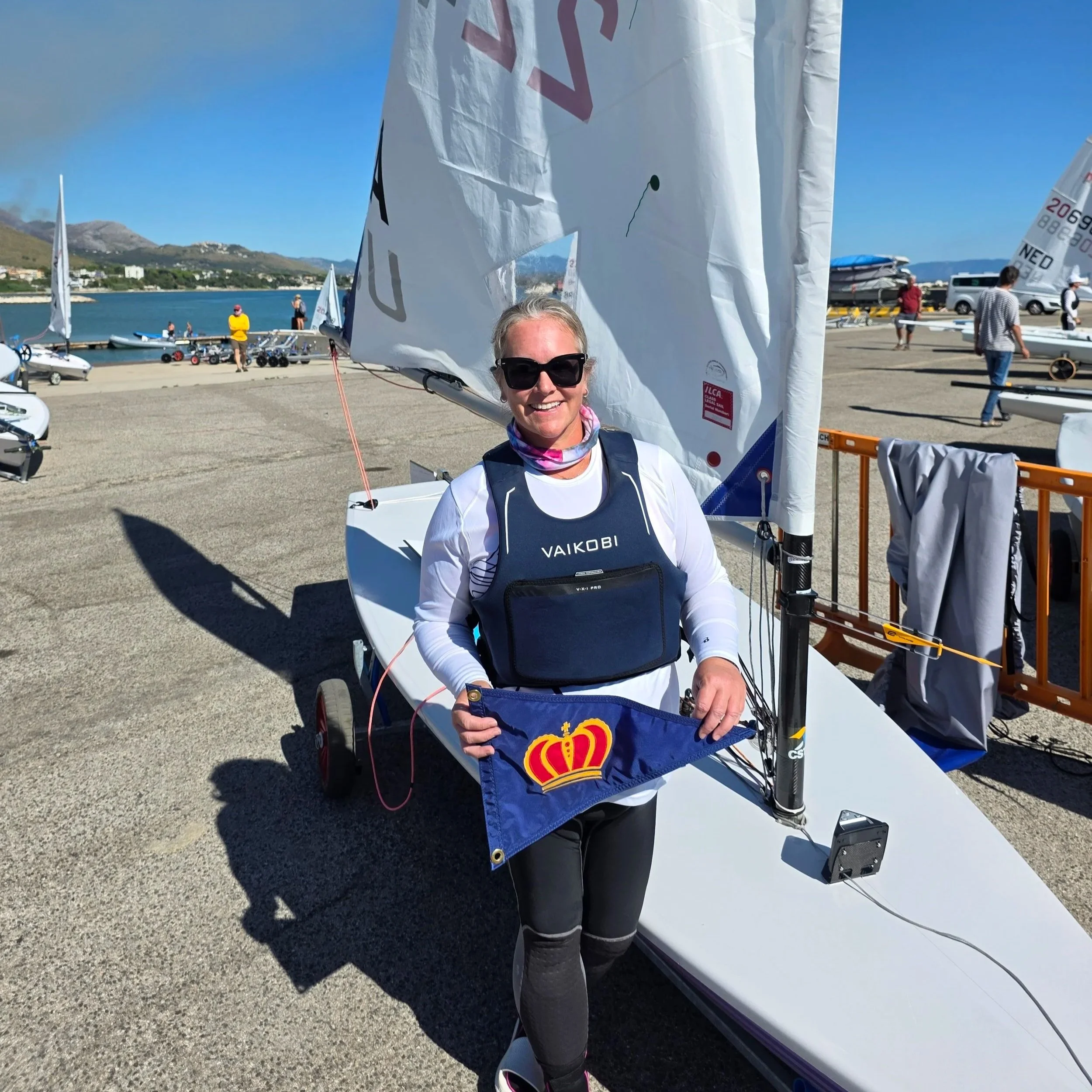 Lauren Wilson stands proudly in front of her ILCA 6 dinghy holding a Coronado Yacht Club burgee, symbolizing her roots and community support.