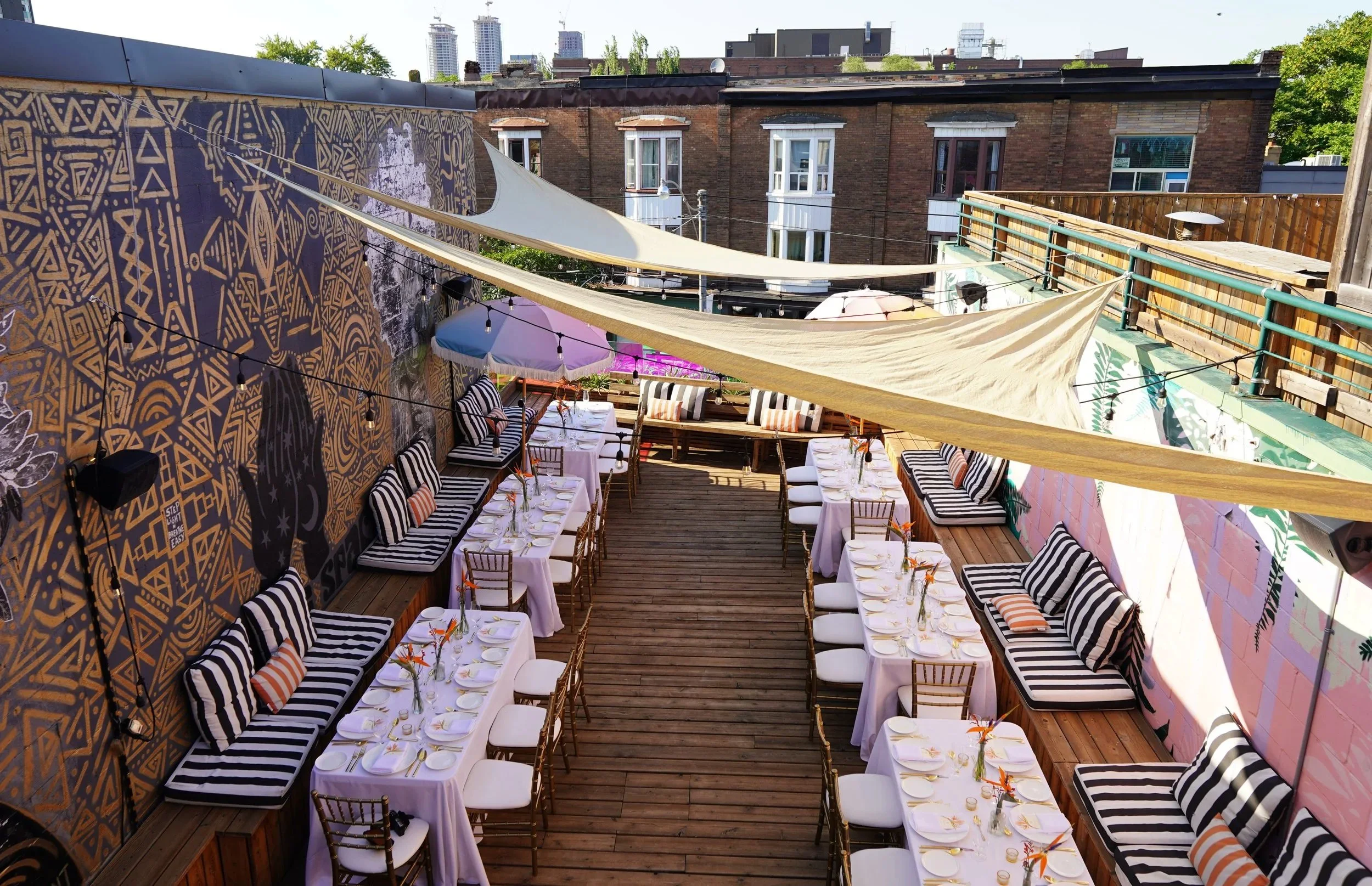 Outdoor rooftop dining area with tables set for a meal, decorated with striped cushions and small floral centerpieces, shaded by fabric sails for outdoor dinner party event at Ode Hotel in Toronto.