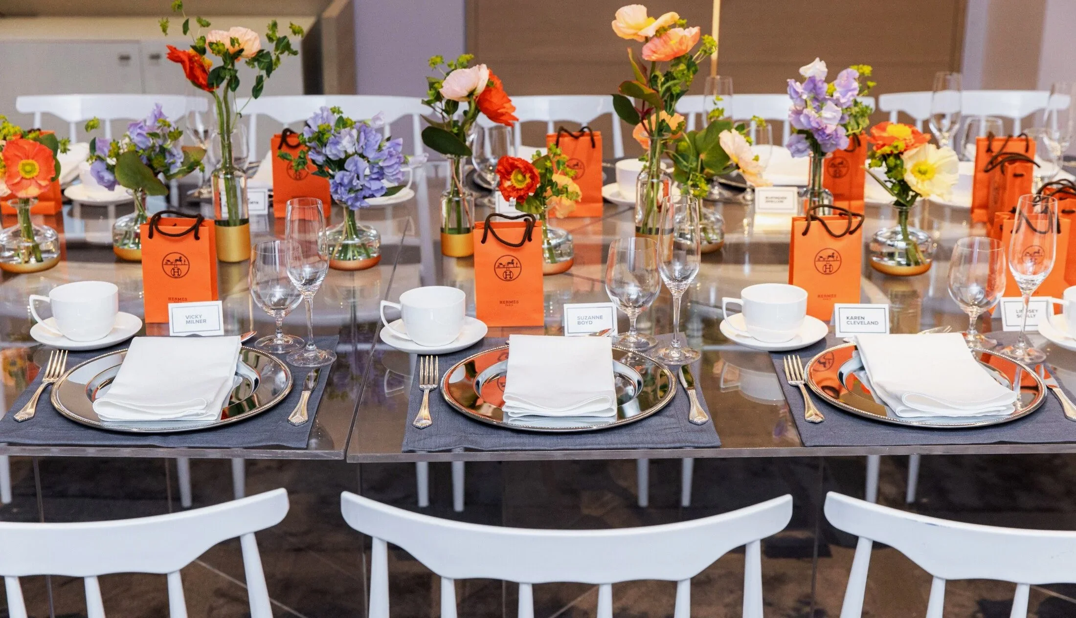 A banquet table decorated with floral centerpieces in vases, orange gift bags, and place settings with silver chargers, white napkins, glasses, and cups for a formal event for The Bay private designer lunch event for International Women's Day.