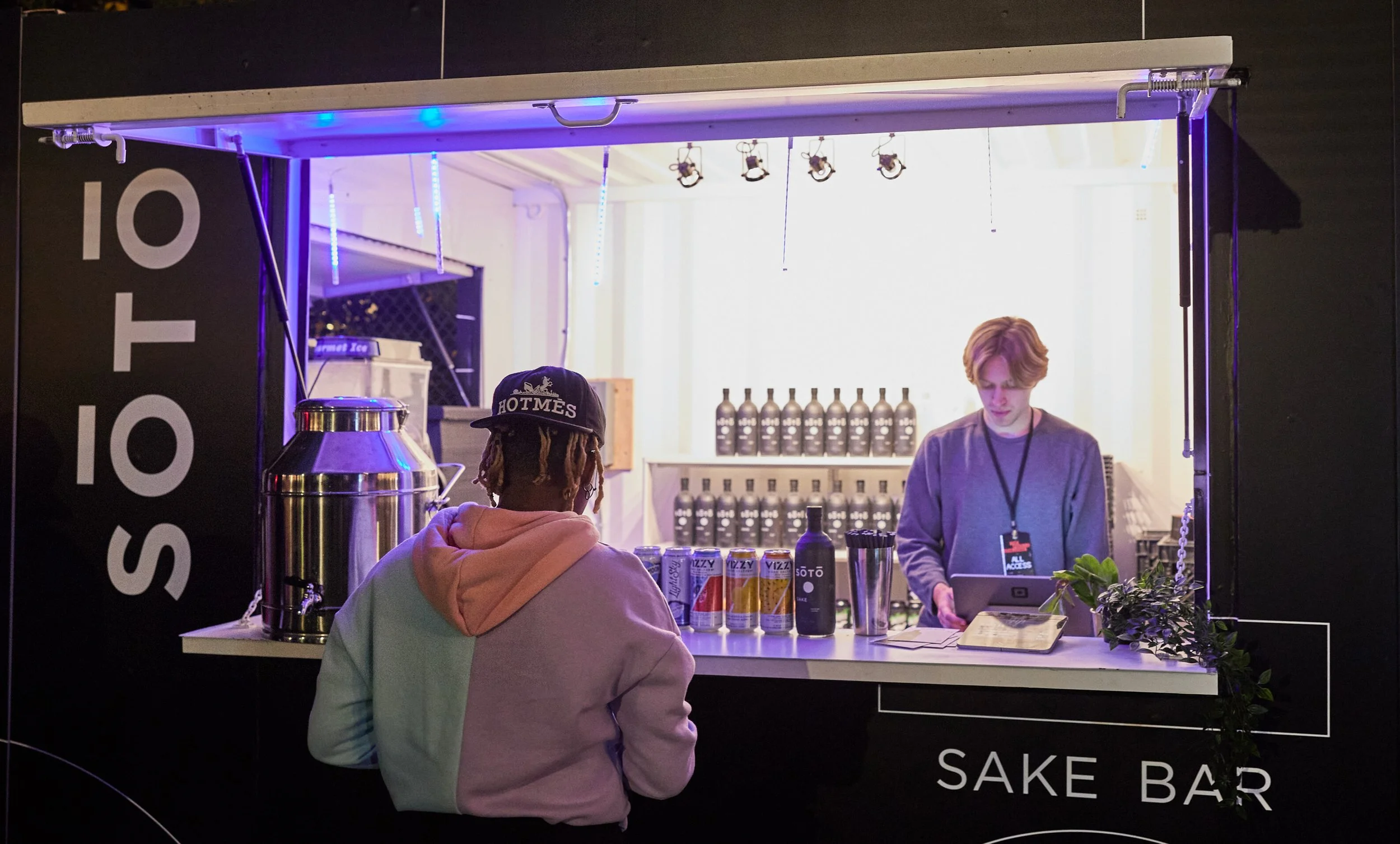 A person facing a sake bar counter with bottles and cans, talking to a bartender behind the counter for an outdoor Toronto event at Stackt Market.