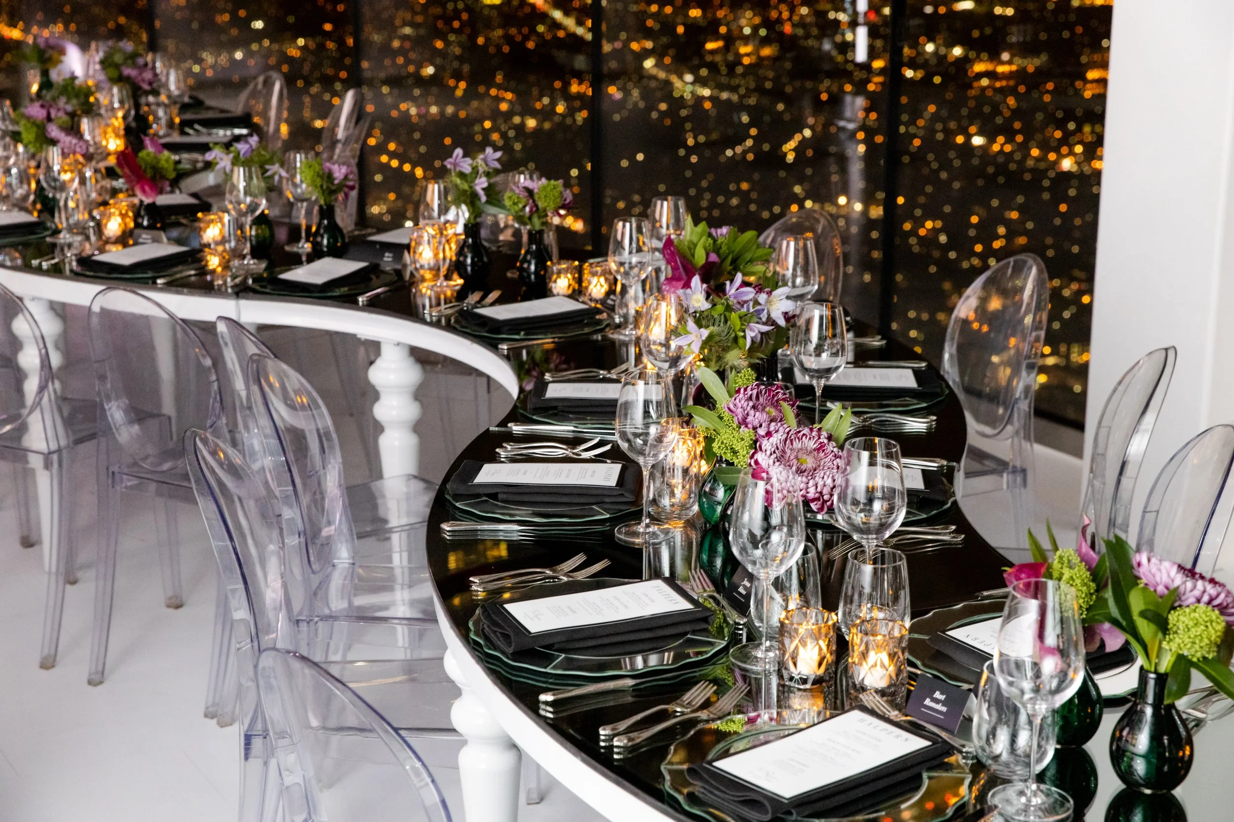 Elegant dining table set with flowers, wine glasses, candles, and black and white menus in a modern indoor setting at night in CN Tower for private fashion designer dinner event.