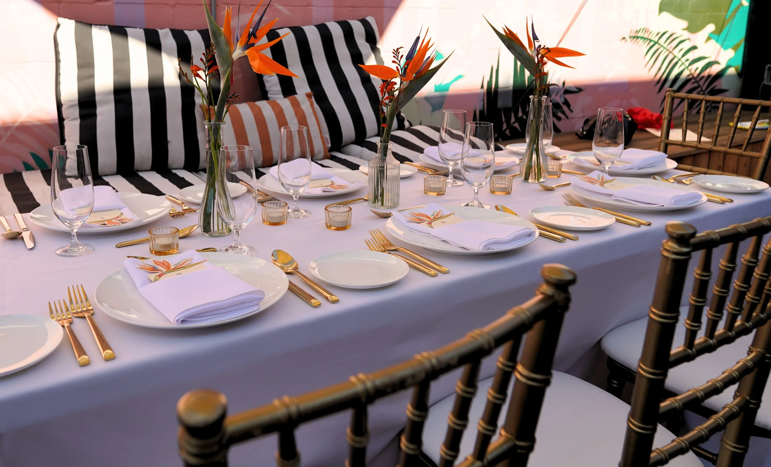 Elegant outdoor dining table decorated with white tablecloth, gold cutlery, white plates with napkins, and tall floral centerpieces featuring orange and green flowers, set for a private dinner party event at Ode Hotel.