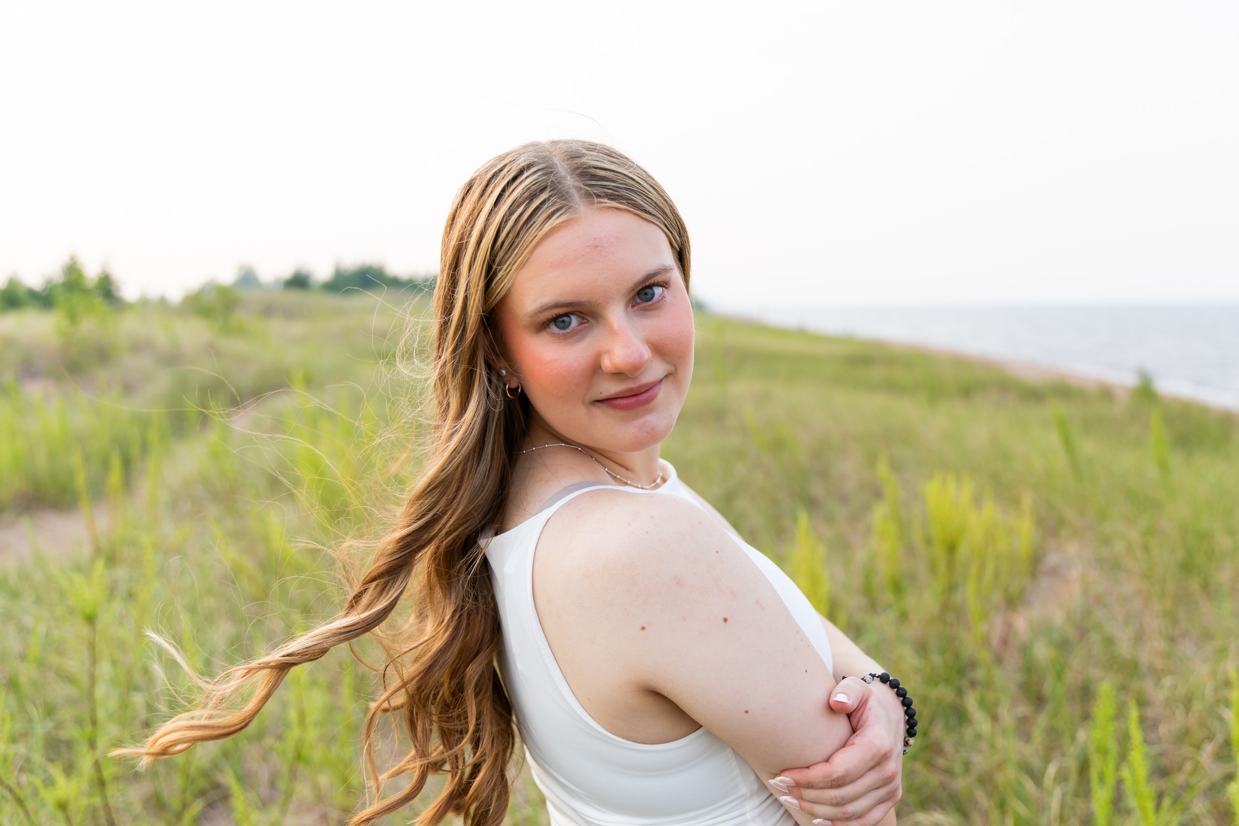 A young woman with long, wavy hair standing outdoors near a grassy area and water under a bright sky, looking at the camera.