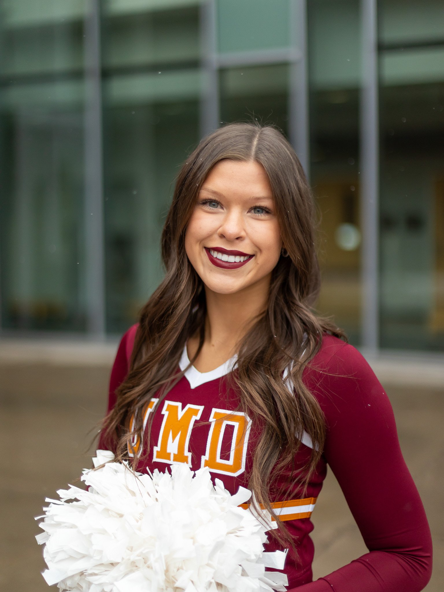 A young woman with long brown hair and blue eyes, smiling, dressed in a maroon cheerleading uniform with white and gold accents, holding white pom-poms, standing outside in front of a modern glass building.