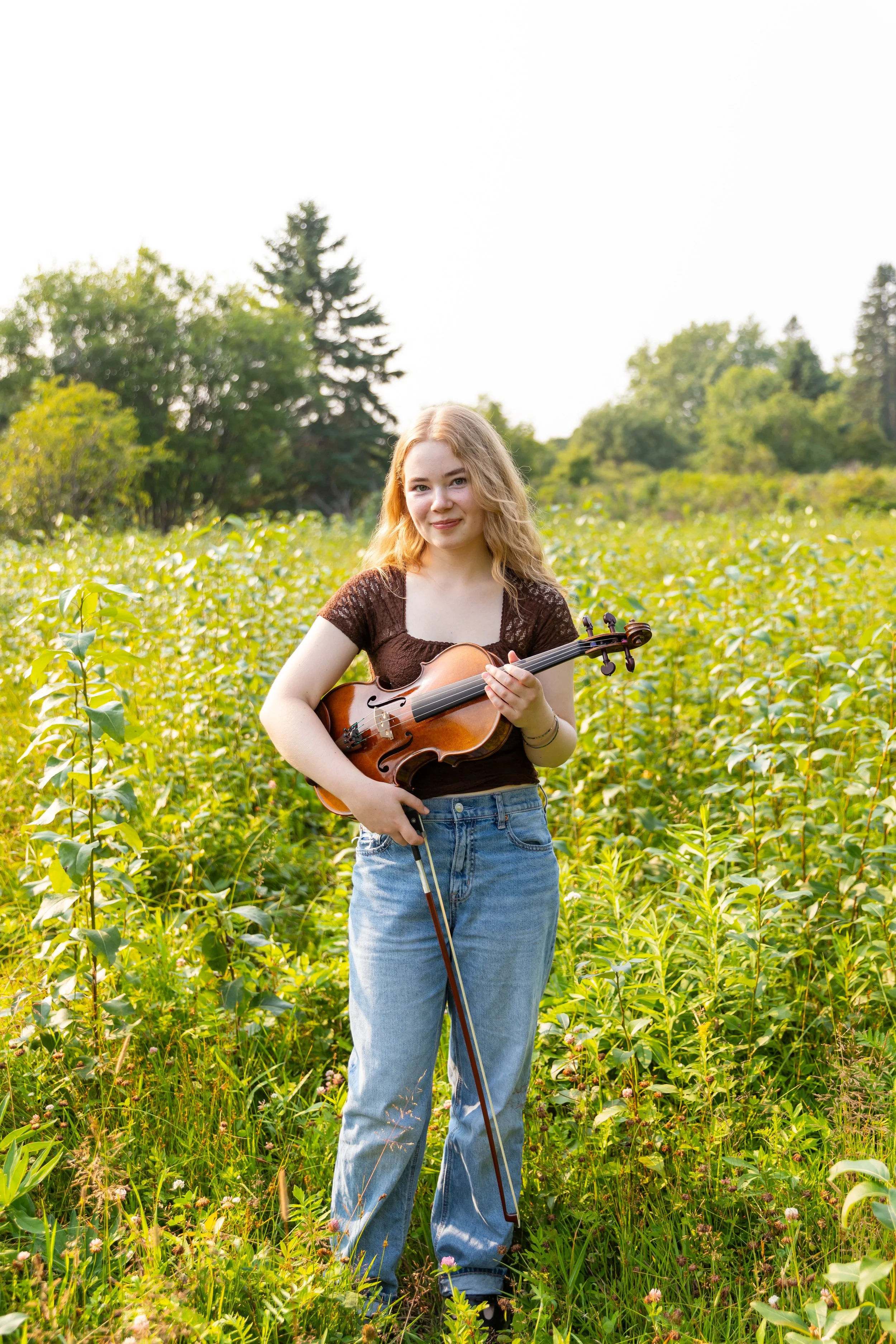 A young woman with long blonde hair and a brown top standing in a green field, holding a violin and bow, with trees in the background.