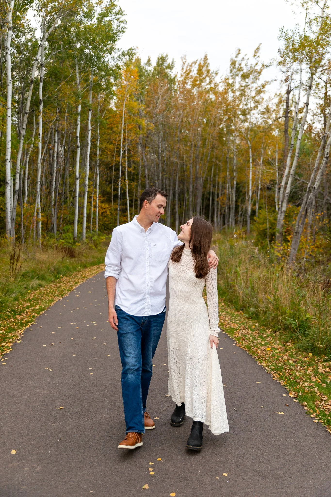 A couple walking arm-in-arm on a paved path through a forest with autumn-colored trees.