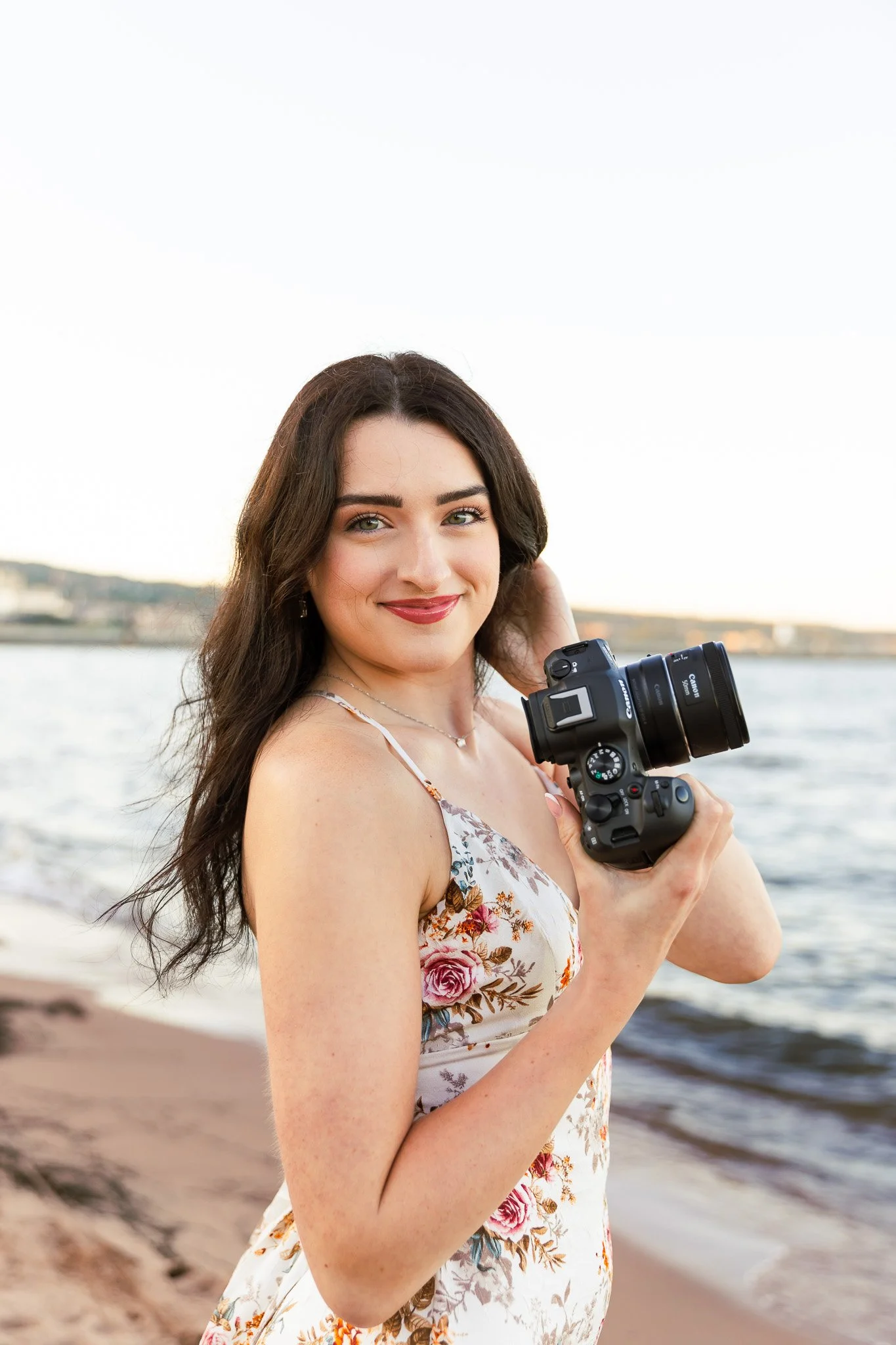 Young woman with long dark hair holding a professional camera by a river or lake during sunset, smiling and wearing a floral dress.