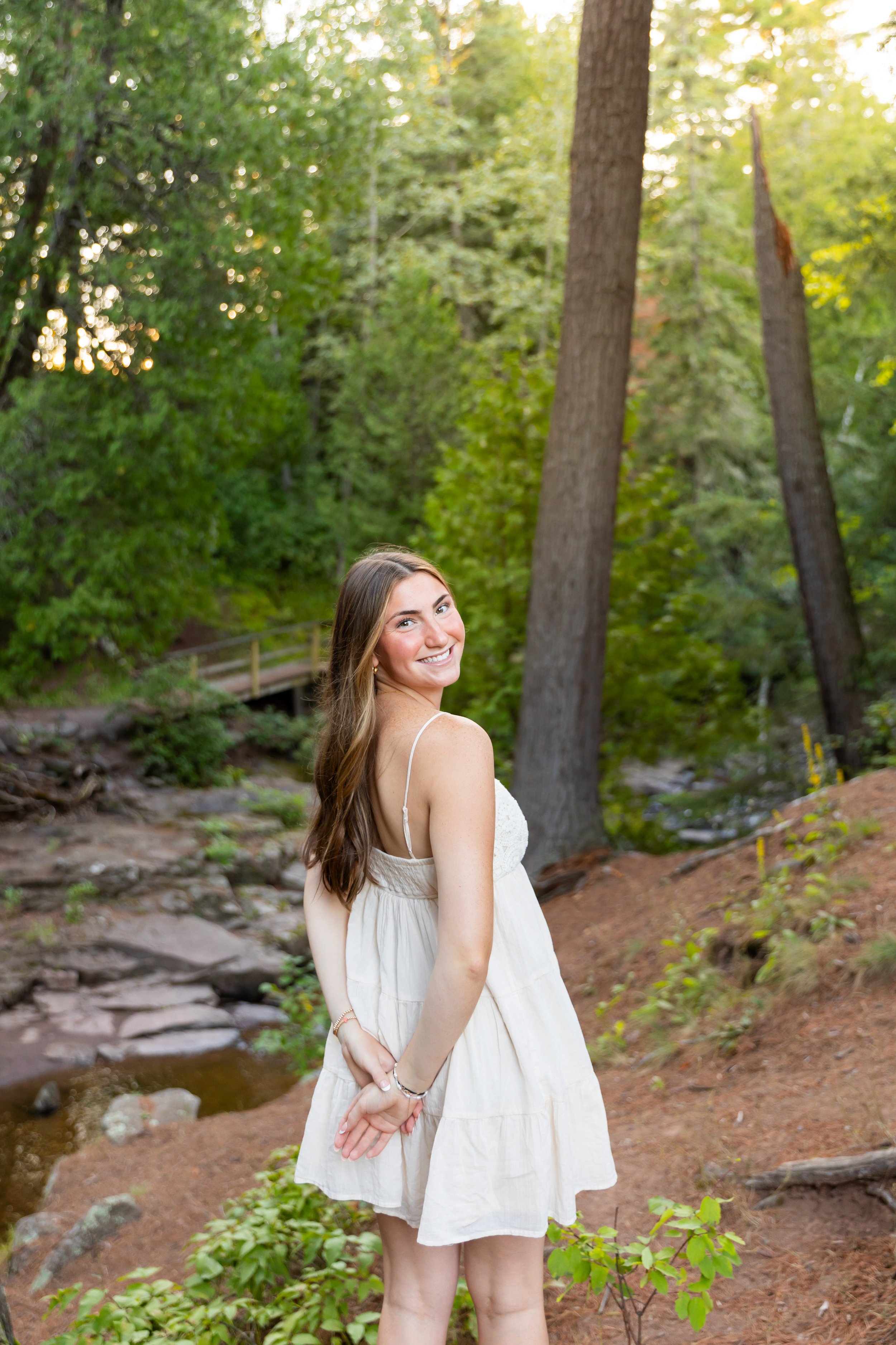 A young woman in a light-colored sundress smiling and looking over her shoulder on a wooded trail with tall trees and a small creek in the background.