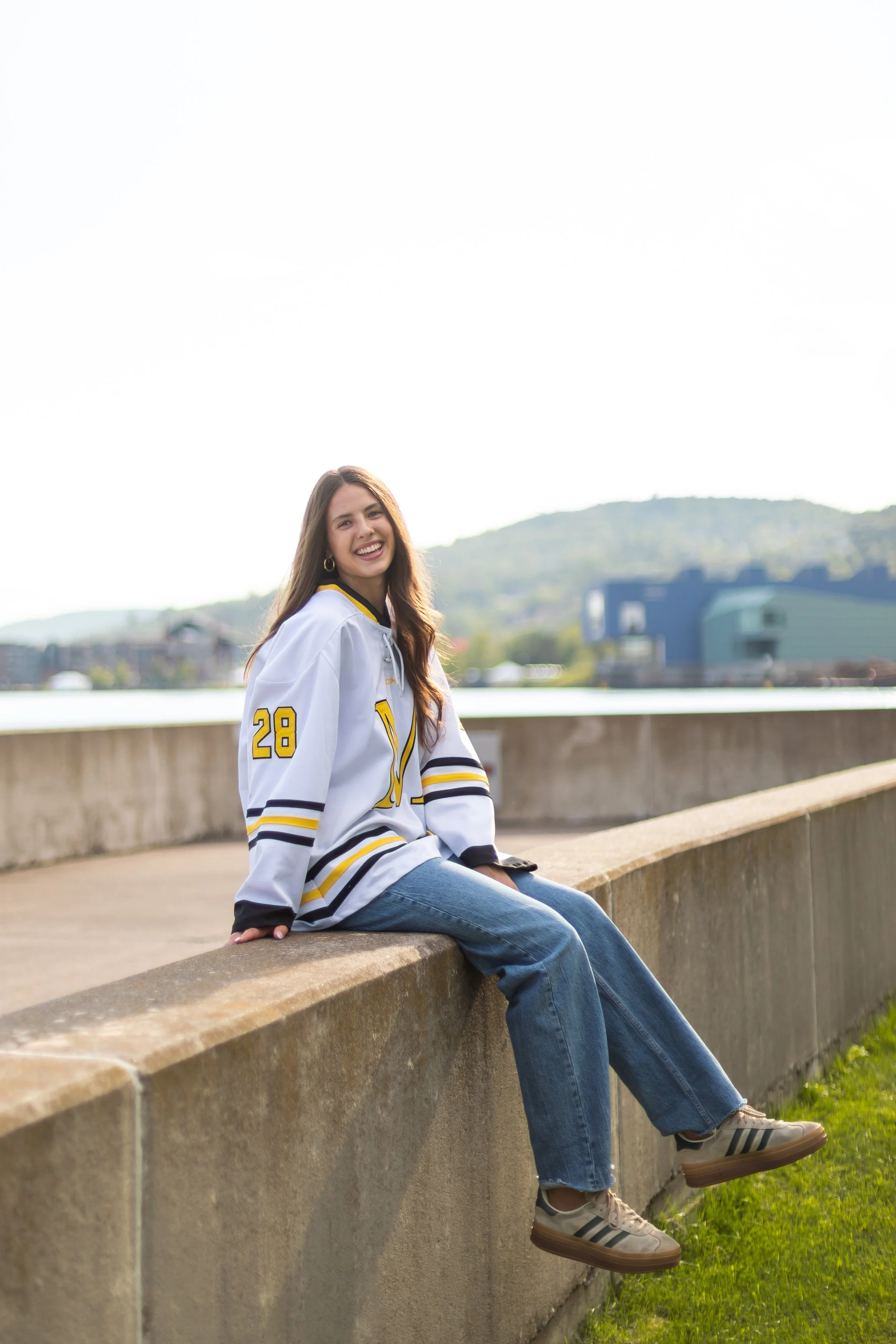 Young woman sitting on a concrete ledge outdoors, smiling, wearing a white hockey jersey with yellow and black details, blue jeans, and beige sneakers, with a scenic background of water and hills.