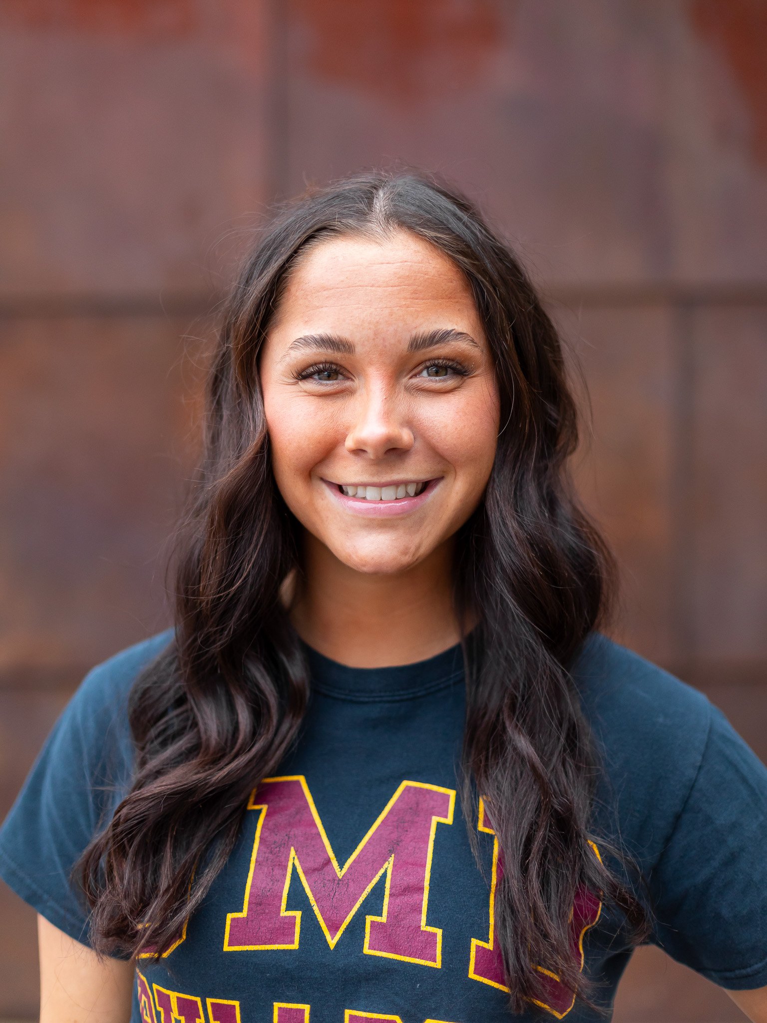 Young woman with long dark hair smiling, wearing a navy t-shirt with a large maroon and yellow letter M, standing outdoors with a brown wall background.