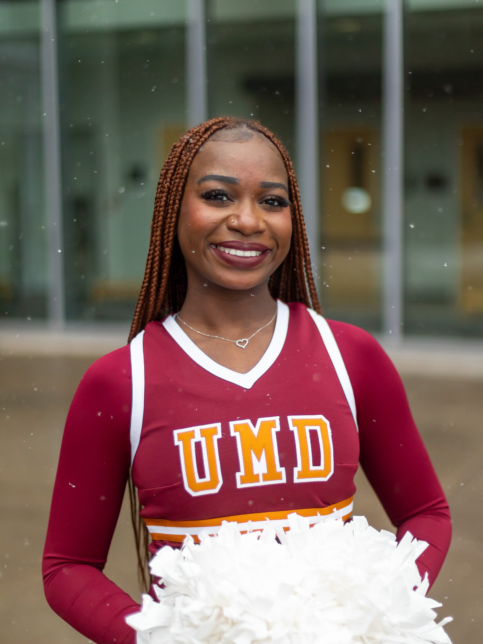 A young woman with braided hair smiling and holding white pom-poms, wearing a maroon UMD cheerleading uniform, standing in front of a glass building.