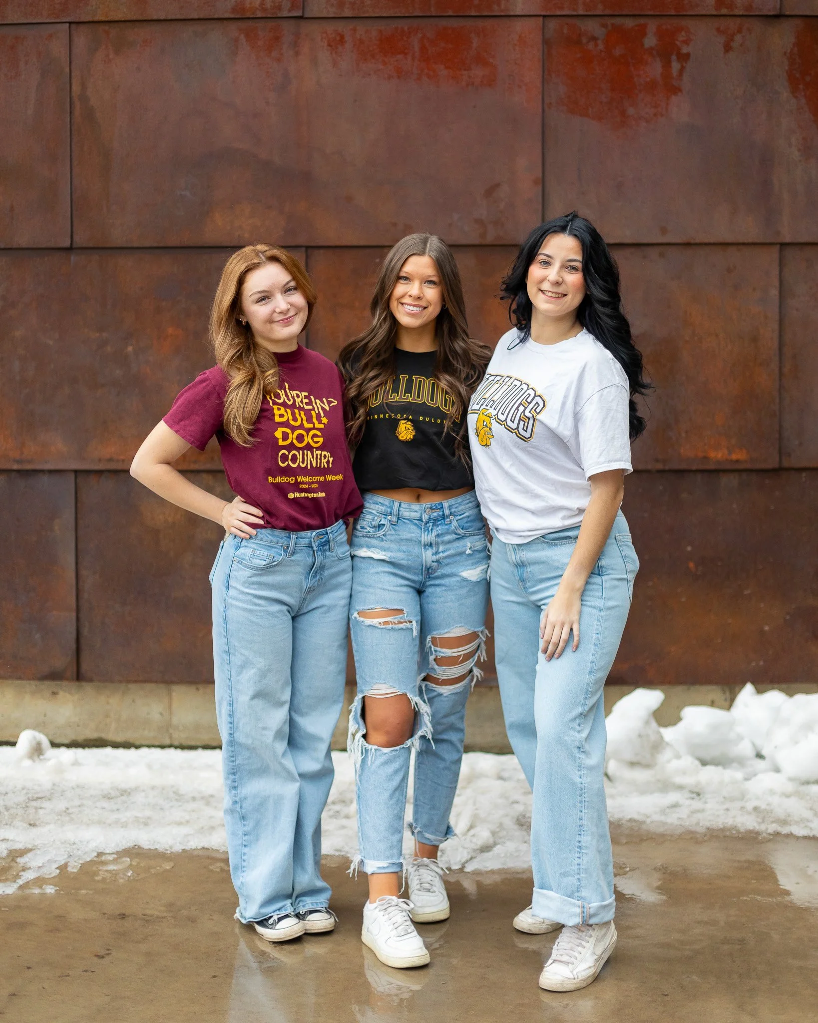 Three young women standing outdoors in front of a rust-colored wall, smiling, wearing casual clothing and Bulldogs-themed shirts, with snow on the ground.