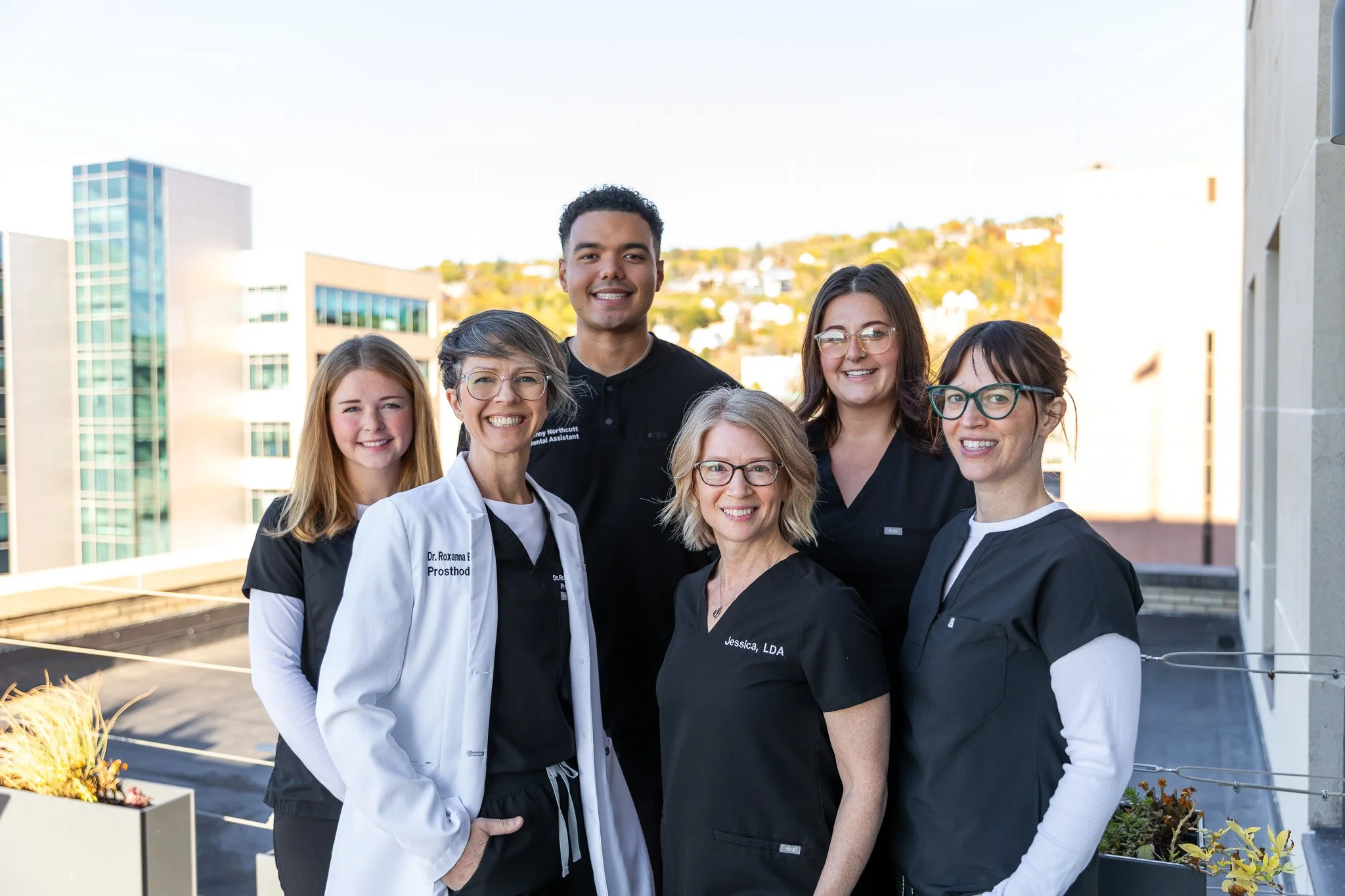 Group of six medical professionals smiling outdoors on a balcony with city buildings in the background.
