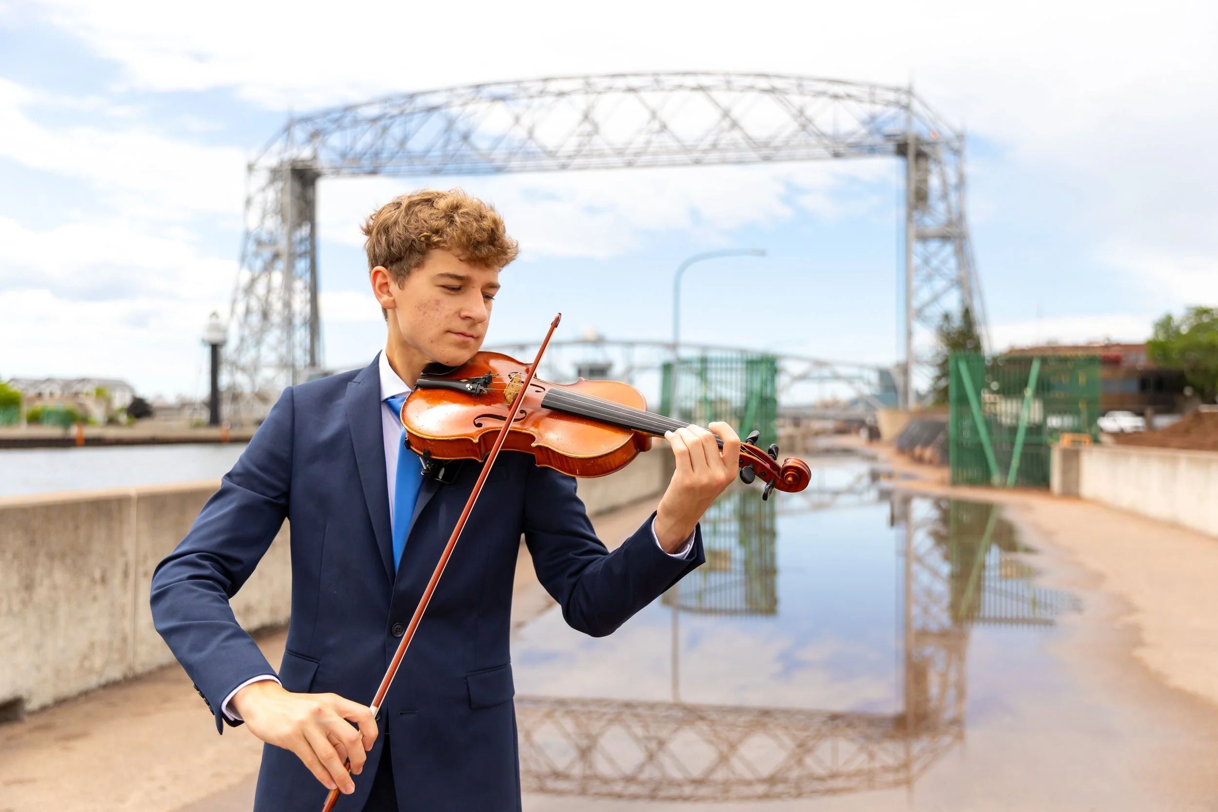 A young man in a suit playing the violin outdoors near a canal with industrial structures in the background.