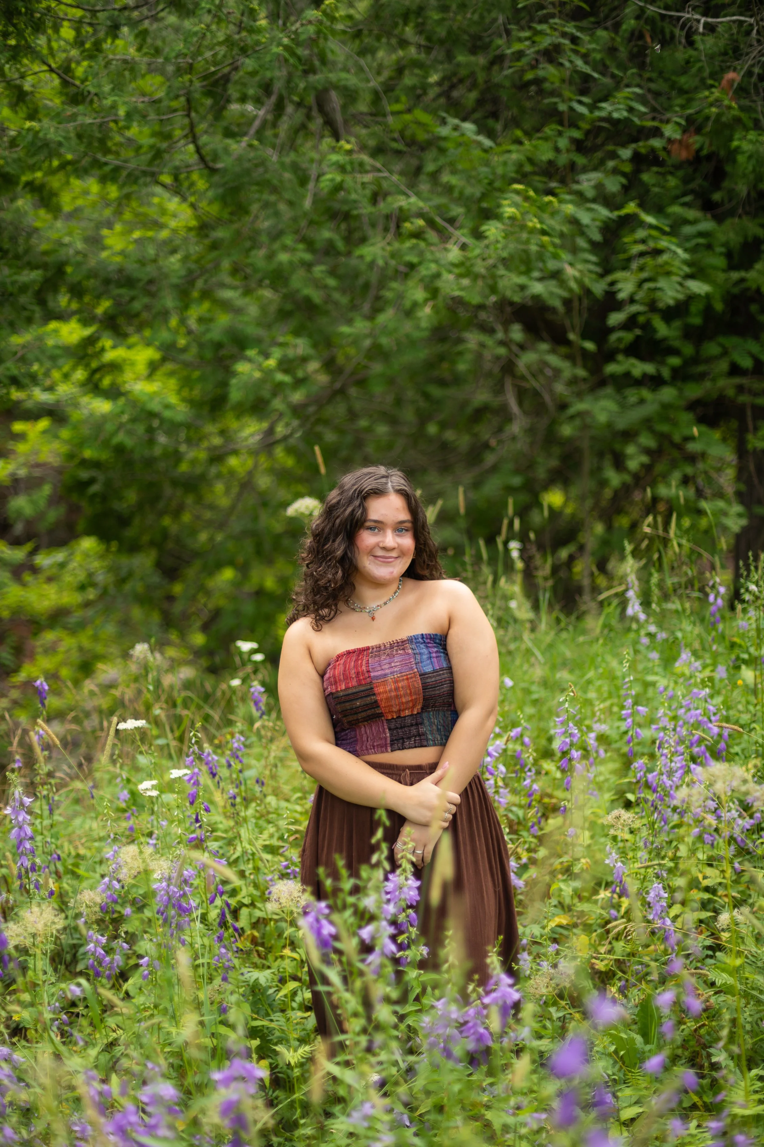 A young woman with curly dark hair wearing a colorful strapless top and a brown skirt, standing amidst purple wildflowers in a lush green outdoor setting.